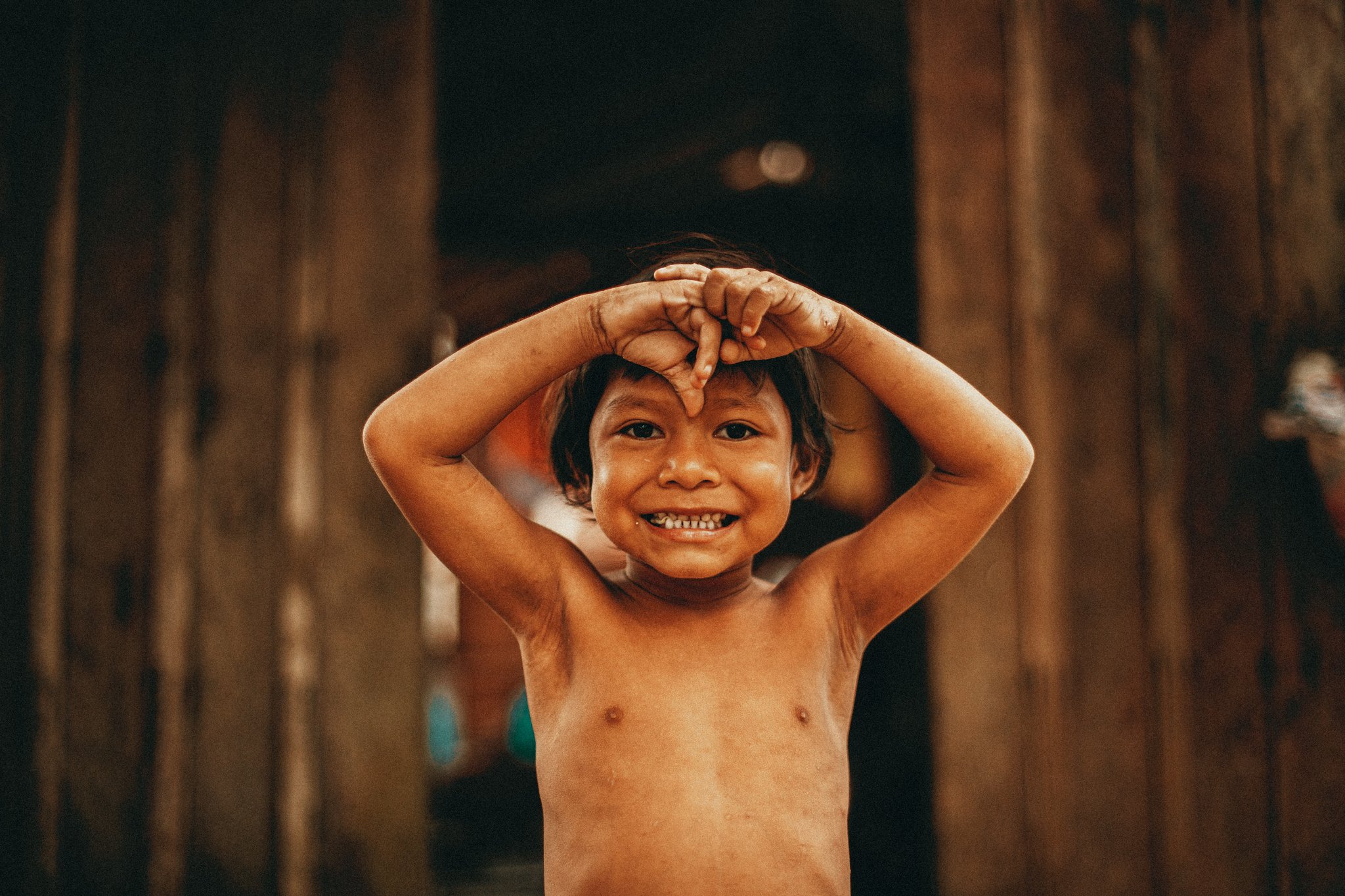 Unscripted joy from a riverside village visit. A laughing child in a riverside community near Anavilhanas Jungle Lodge Brazil, hands raised playfully above their head in front of a wooden wall