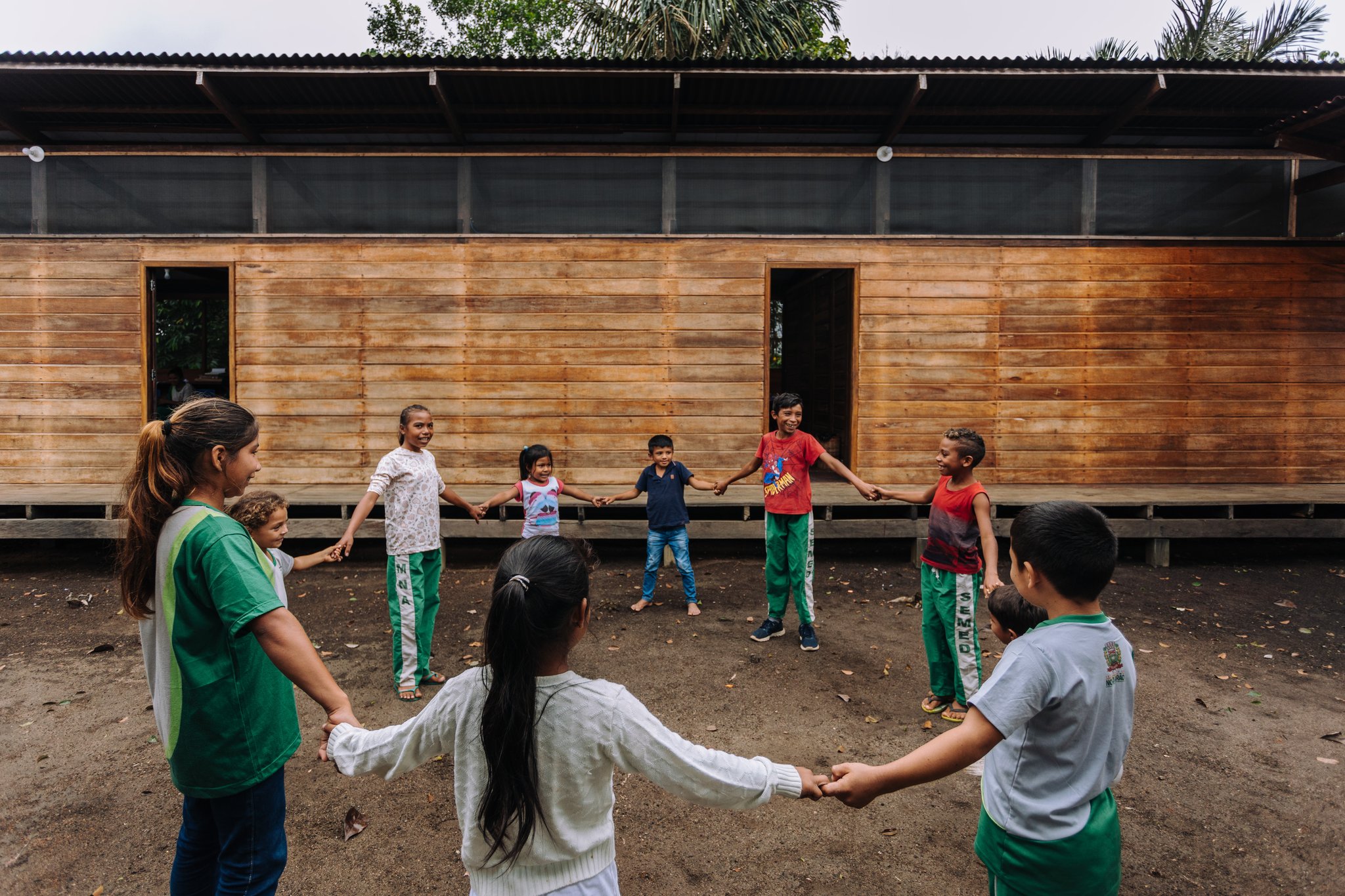 Laughter in a circle — community life at its most joyful. Children holding hands in a circle and playing outside a wooden community building during a riverside village visit at Anavilhanas Jungle Lodge Brazil