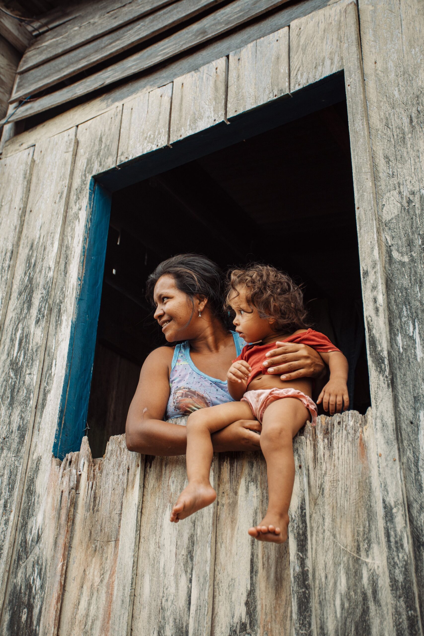 A mother, a toddler, a window — everyday life along the river. A smiling woman holds a barefoot toddler on the sill of a weathered wooden window during a riverside community visit near Anavilhanas Jungle Lodge Brazil