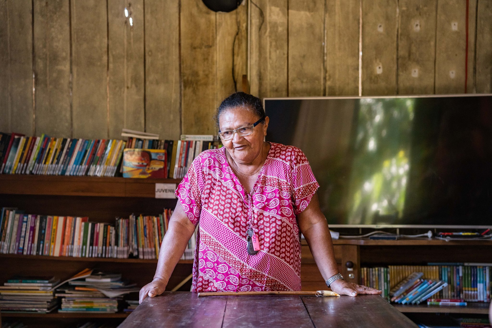 Knowledge keeper: a community librarian shares her world with visitors. Photo: Alex Fisberg A local woman in a pink patterned dress stands at a wooden table inside a community library near Anavilhanas Jungle Lodge Brazil, with colourful bookshelves lining the timber walls behind her