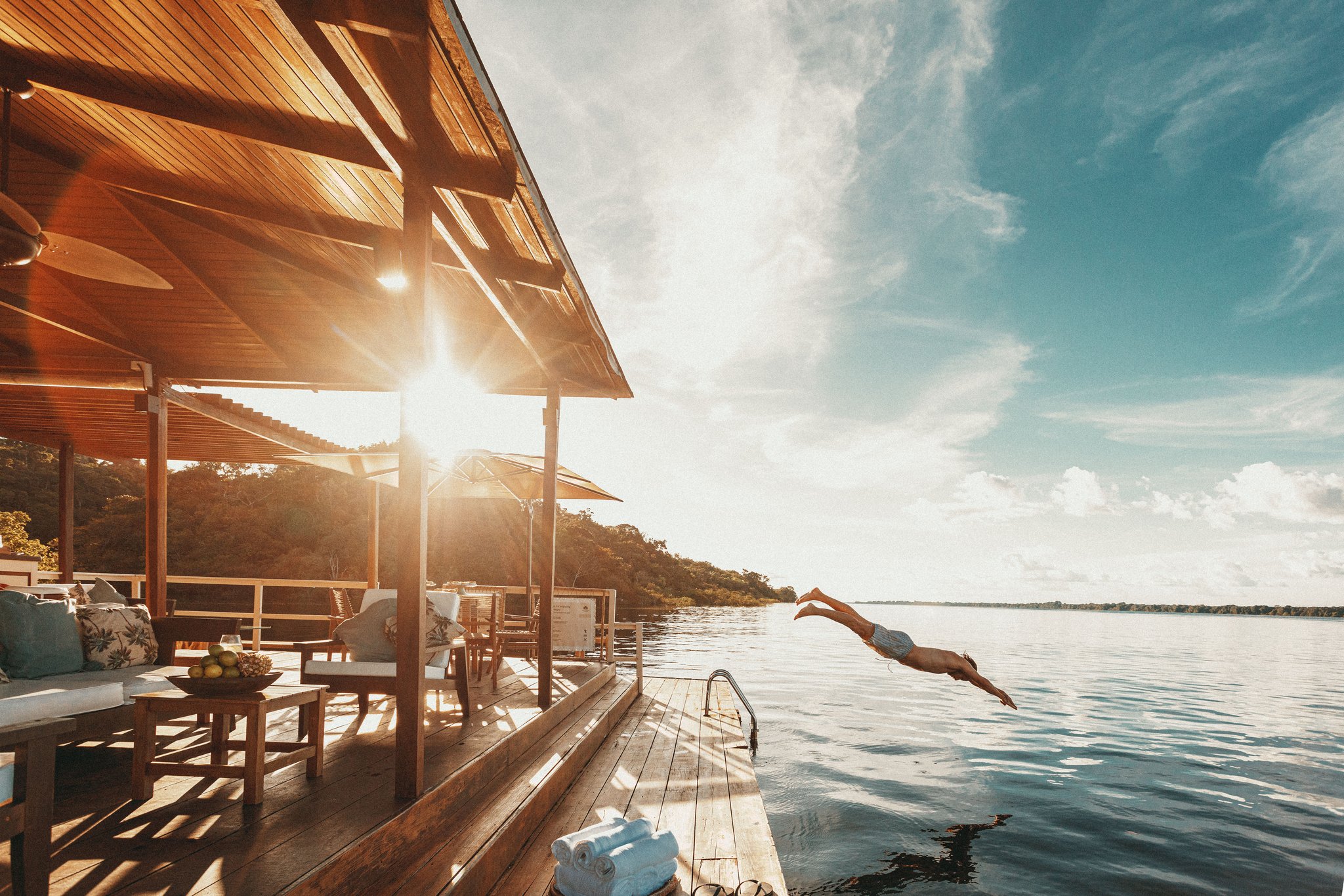 Take the plunge — straight from the deck into the Rio Negro. A guest dives from the wooden deck of Anavilhanas Jungle Lodge Brazil into the calm Rio Negro at golden hour, with a furnished open-air lounge and lush forest visible in the background