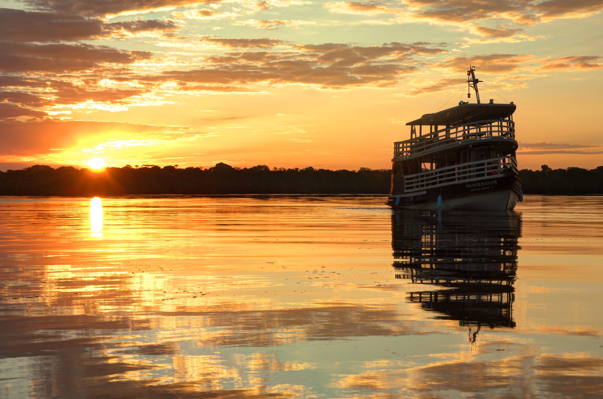 Sunset burns across the Rio Negro — the day's last light, absolutely still. The Anavilhanas Jungle Lodge Brazil expedition boat moored on the glassy Rio Negro beneath a blazing orange sunset, its reflection perfectly mirrored on the still water
