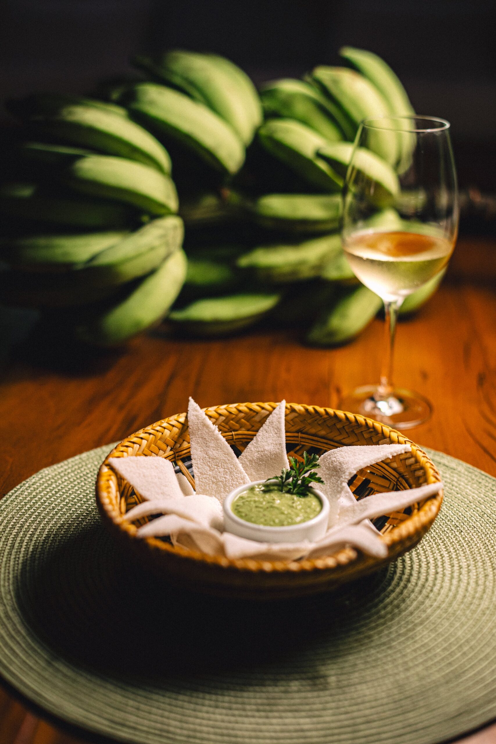 Local ingredients, plated with quiet elegance. A woven rattan basket holding white flatbread pieces arranged around a small bowl of green herb dip, served at Anavilhanas Jungle Lodge Brazil alongside a glass of white wine, with a bunch of green bananas softly blurred in the background