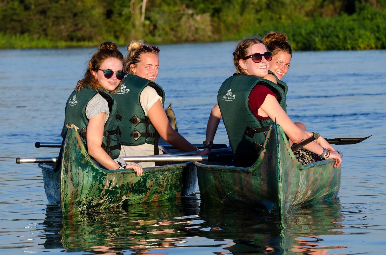 Four smiling women in green life jackets paddling a canoe on a calm river during a budget Pantanal tour, lush green vegetation lining the far bank