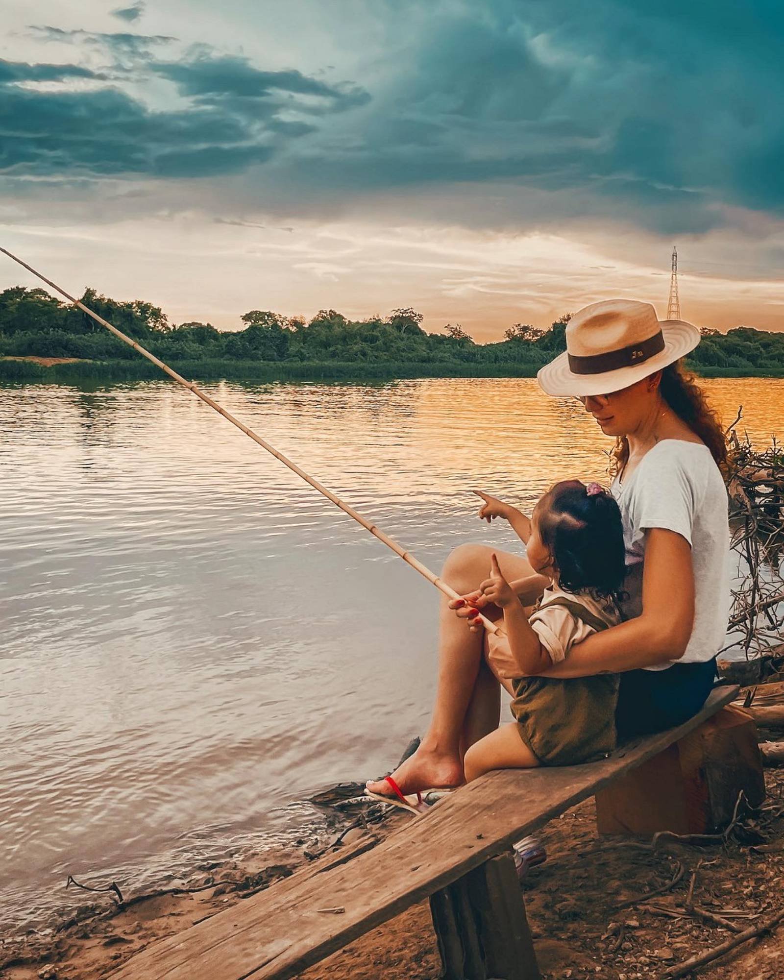 A woman in a panama hat holds a young child on her lap while fishing with a bamboo rod on a calm river at sunset during a budget Pantanal tour, golden light reflecting off the water