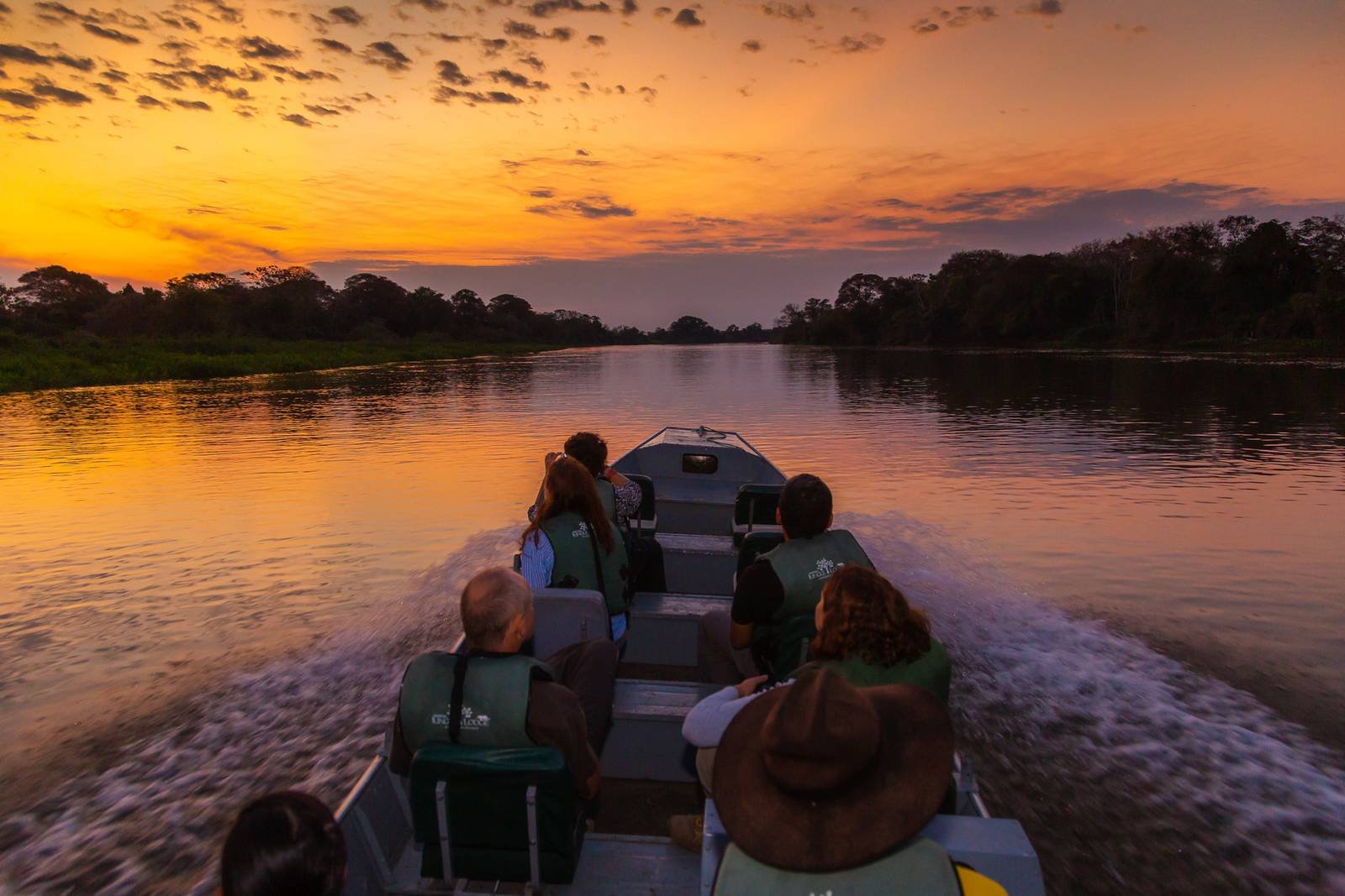Travellers in life jackets aboard a motorboat on a calm river at sunset during a budget Pantanal tour, with a vivid orange sky reflecting on the water and silhouetted jungle on both banks