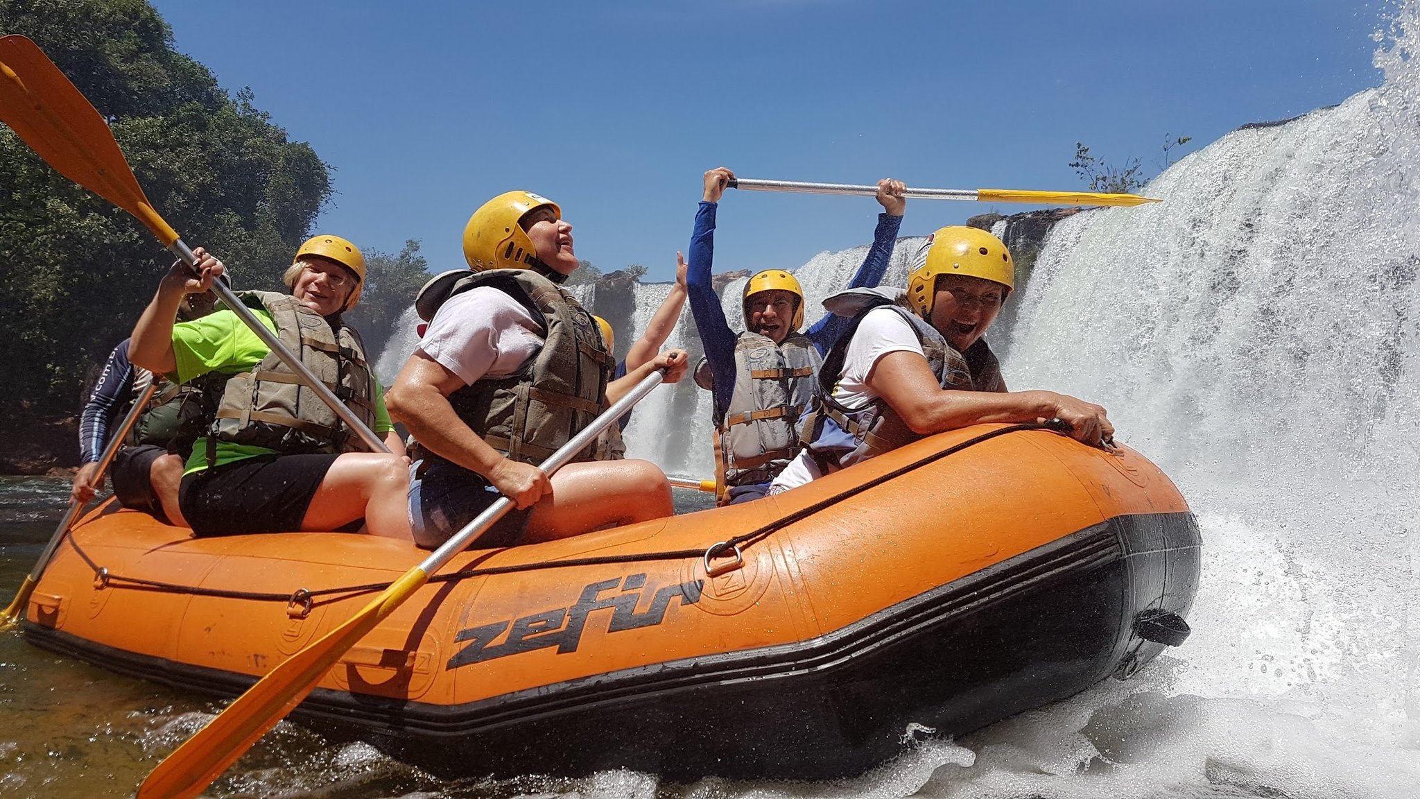 Five excited rafters in yellow helmets and life jackets paddle an orange inflatable raft alongside a thundering waterfall in the Cerrado Brazil wilderness
