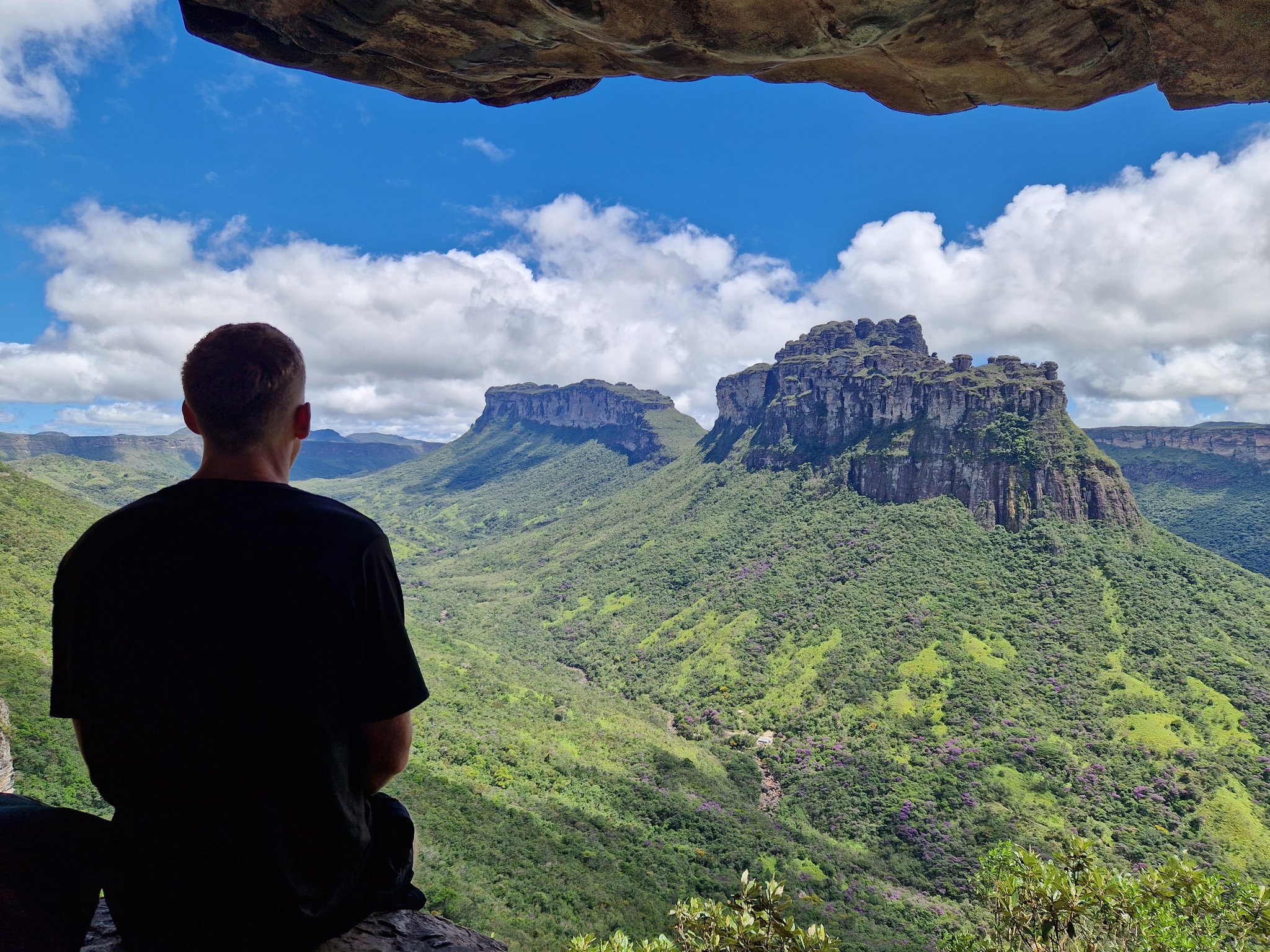 Traveler seated inside a rock shelter gazes out at the lush green valleys and dramatic sandstone mesas of Chapada Diamantina tours landscape under a blue sky