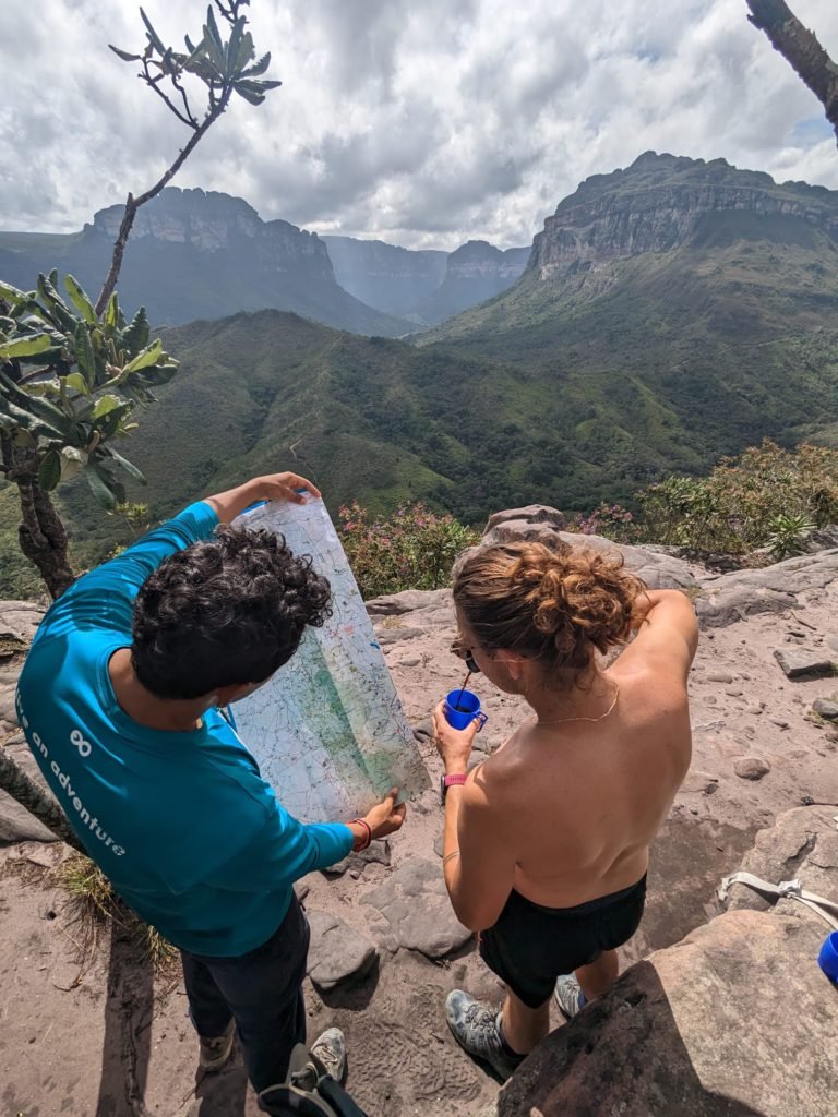 A guide in a blue shirt and a traveler study a trail map together on a rocky outcrop overlooking the green canyon valleys and table-top mountains of Chapada Diamantina National Park