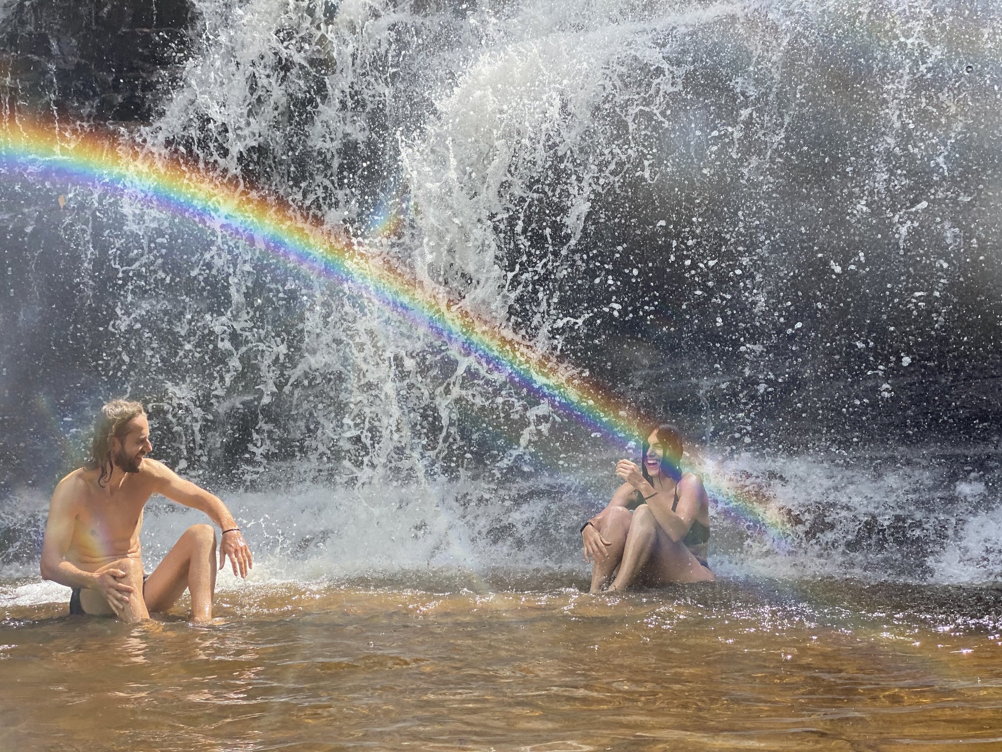 Two travelers sitting and laughing in a shallow waterfall pool on a Chapada Diamantina tour, with a vivid rainbow arcing through the cascading white water behind them