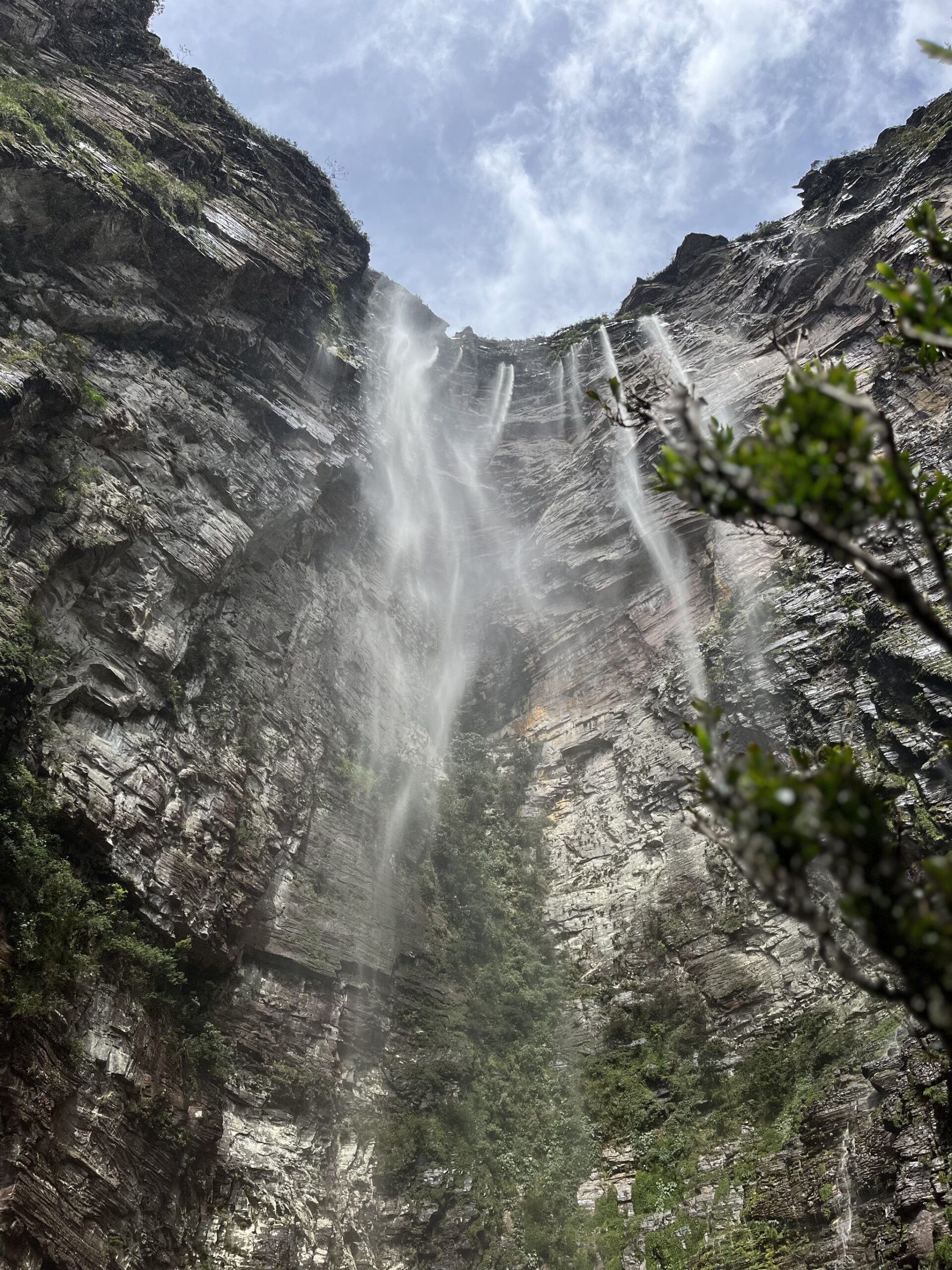 Mist on your face, a wall of rock above — Cachoeirão from below is pure awe. Photo: Lucas Neves Looking straight up at Cachoeirão from its base during the Chapada Diamantina trek, multiple ribbons of white water streaming down a towering vertical sandstone cliff framed by green shrubs and an open sky