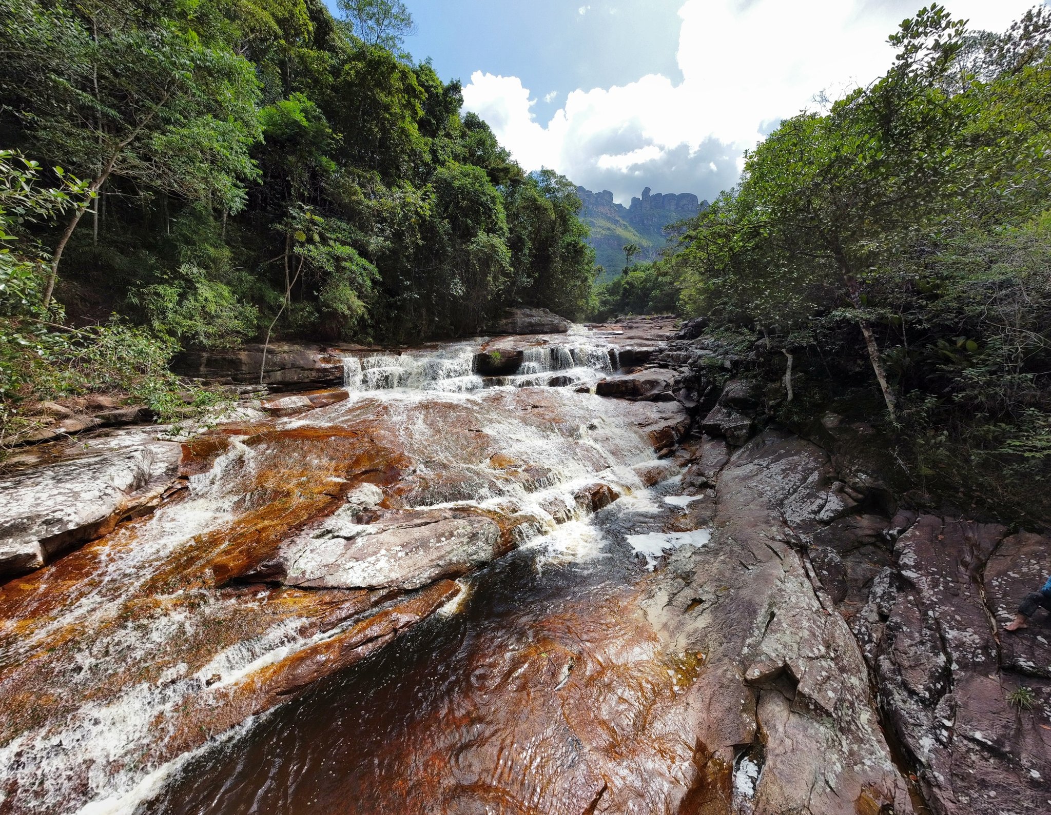Rust-red rock, white rapids, ancient mountains — the Pati River in full flow. White water tumbling over rust-coloured sandstone slabs on a Chapada Diamantina trek, with lush rainforest and distant rocky peaks rising under a cloudy sky