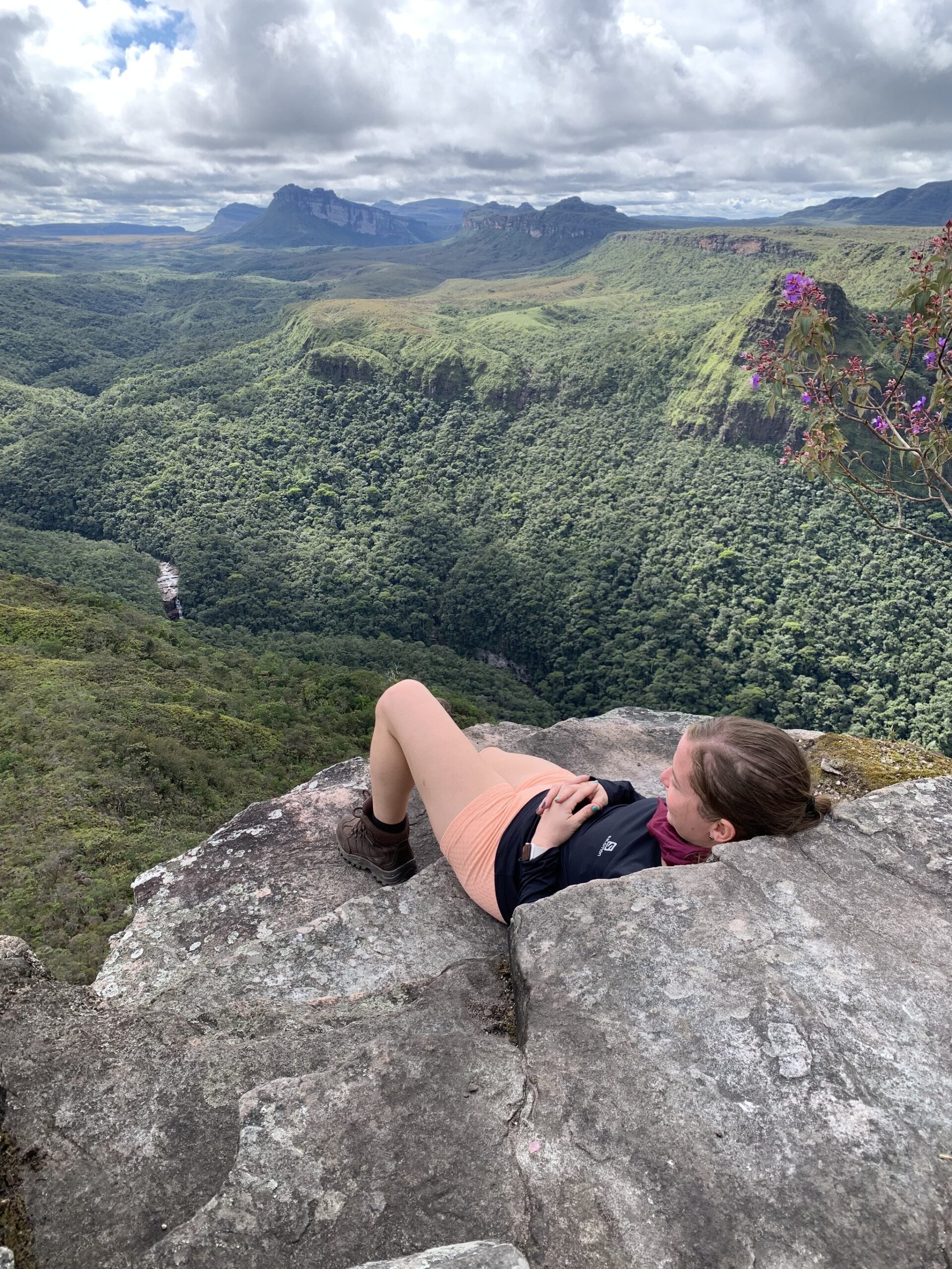 Flat on rock, the whole canyon yours. A hiker lies on a rocky clifftop ledge on a Chapada Diamantina trek, gazing out over a vast green canyon with flat-topped sandstone mesas and a distant waterfall under a cloudy sky