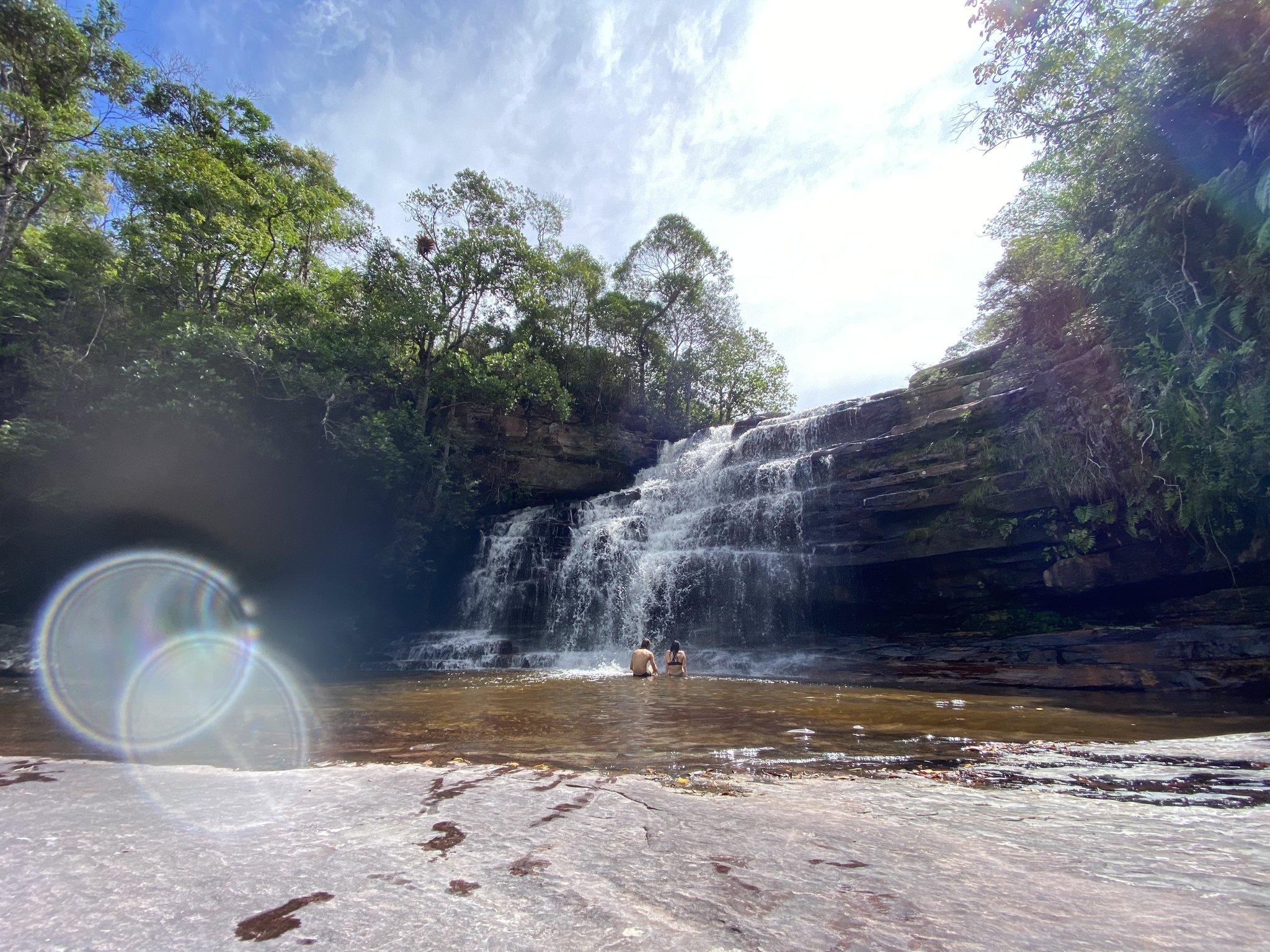 Cool off beneath Cachoeira dos Funis — sandstone, cascading tiers, and pure Chapada magic. Two swimmers wade into the clear natural pool beneath a wide tiered waterfall cascading over sandstone ledges on the Chapada Diamantina trek, surrounded by dense green forest under a bright midday sky