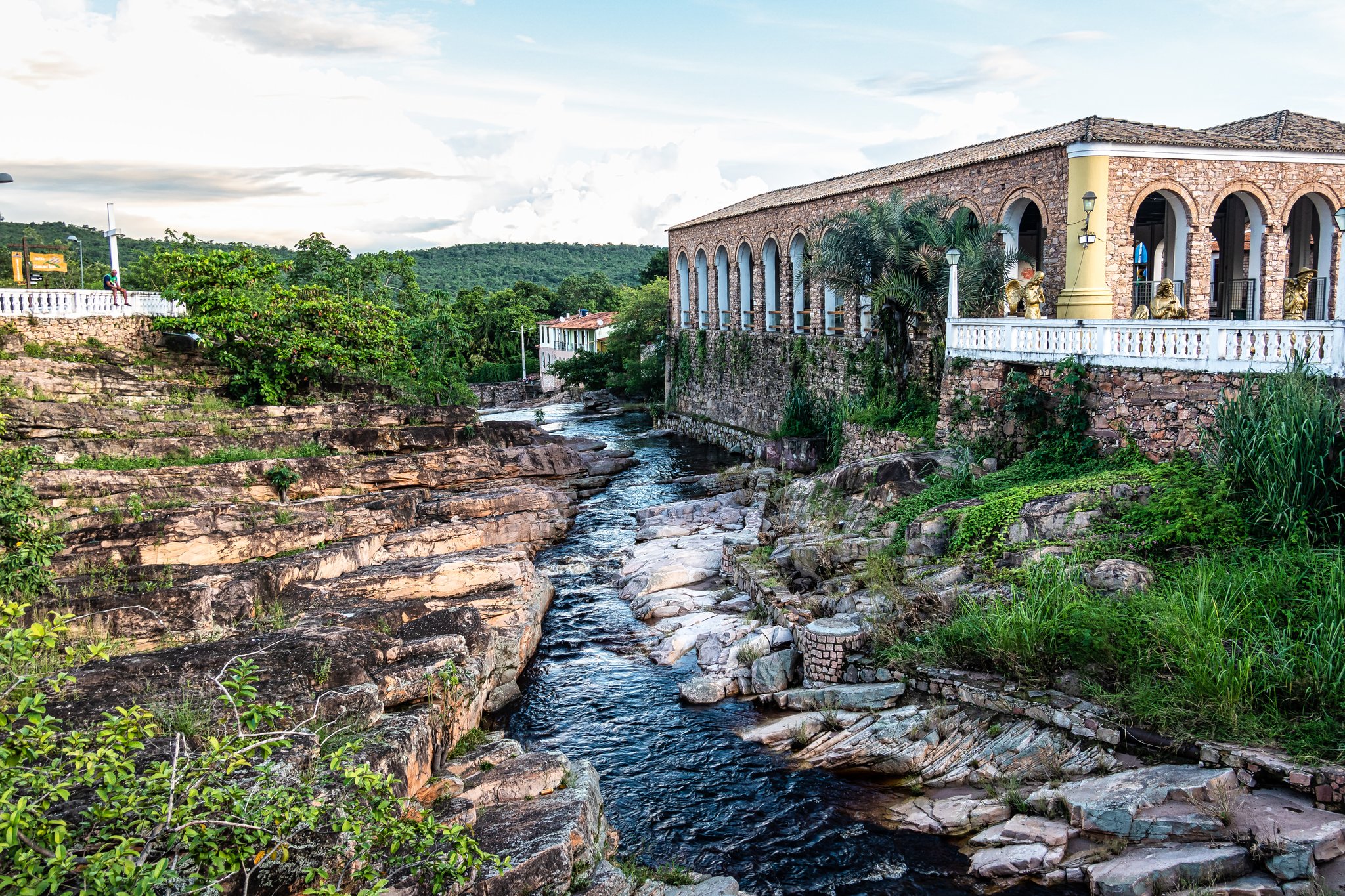 A clear river cuts through layered sandstone rock formations alongside a colonial-era stone building with arched galleries and a white balustrade, surrounded by green hills on a Chapada Diamantina trek gateway town