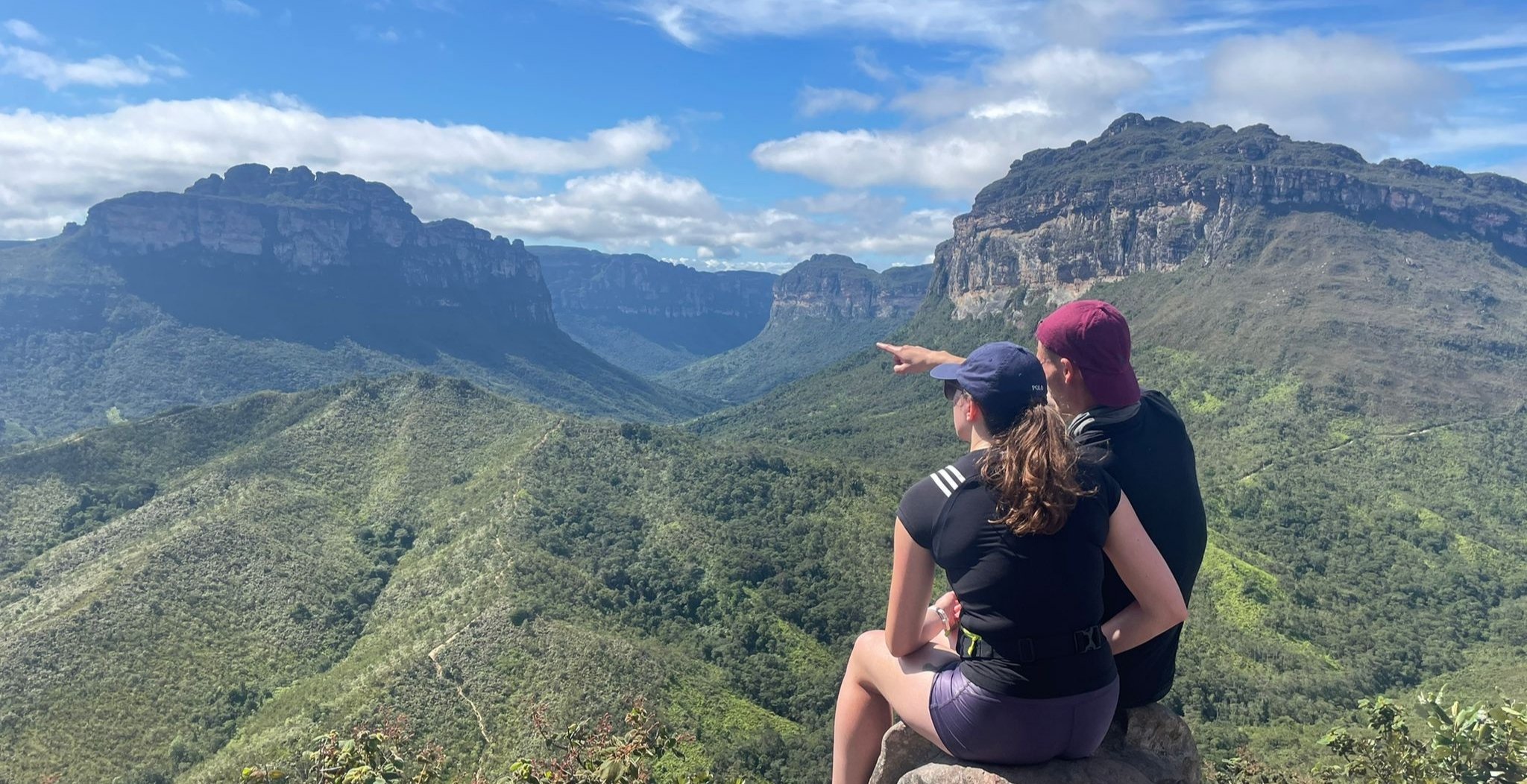 Two hikers sitting on a rock at a high viewpoint on the Chapada Diamantina trek, one pointing across a sweeping green valley framed by towering sandstone mesa cliffs under a blue sky