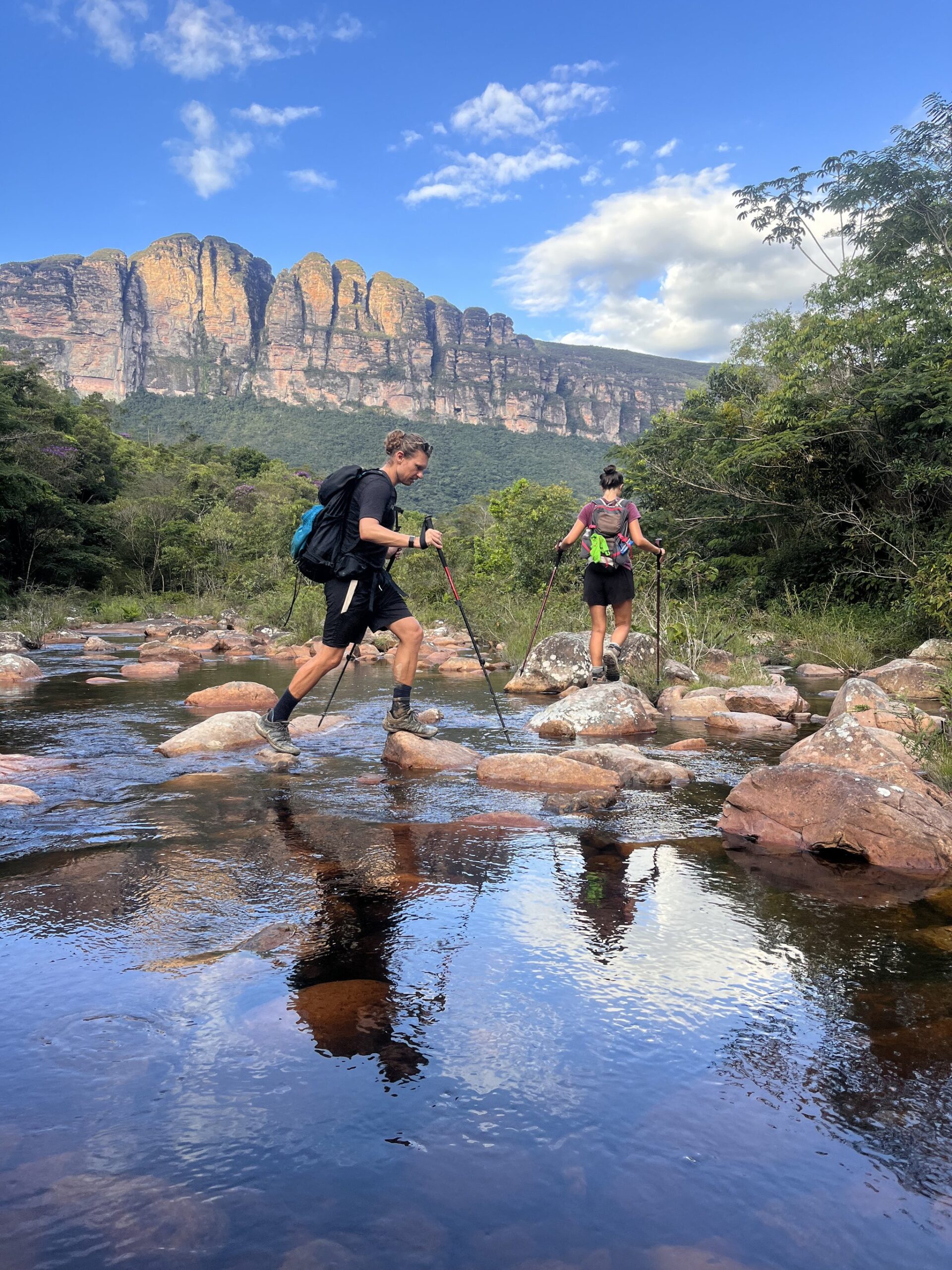 Rock by rock, the river crossed — ancient cliffs watching every step. Photo: Lucas Neves Two hikers with trekking poles stepping across river rocks on a Chapada Diamantina trek, with dramatic sandstone escarpments rising behind lush green vegetation under a blue sky