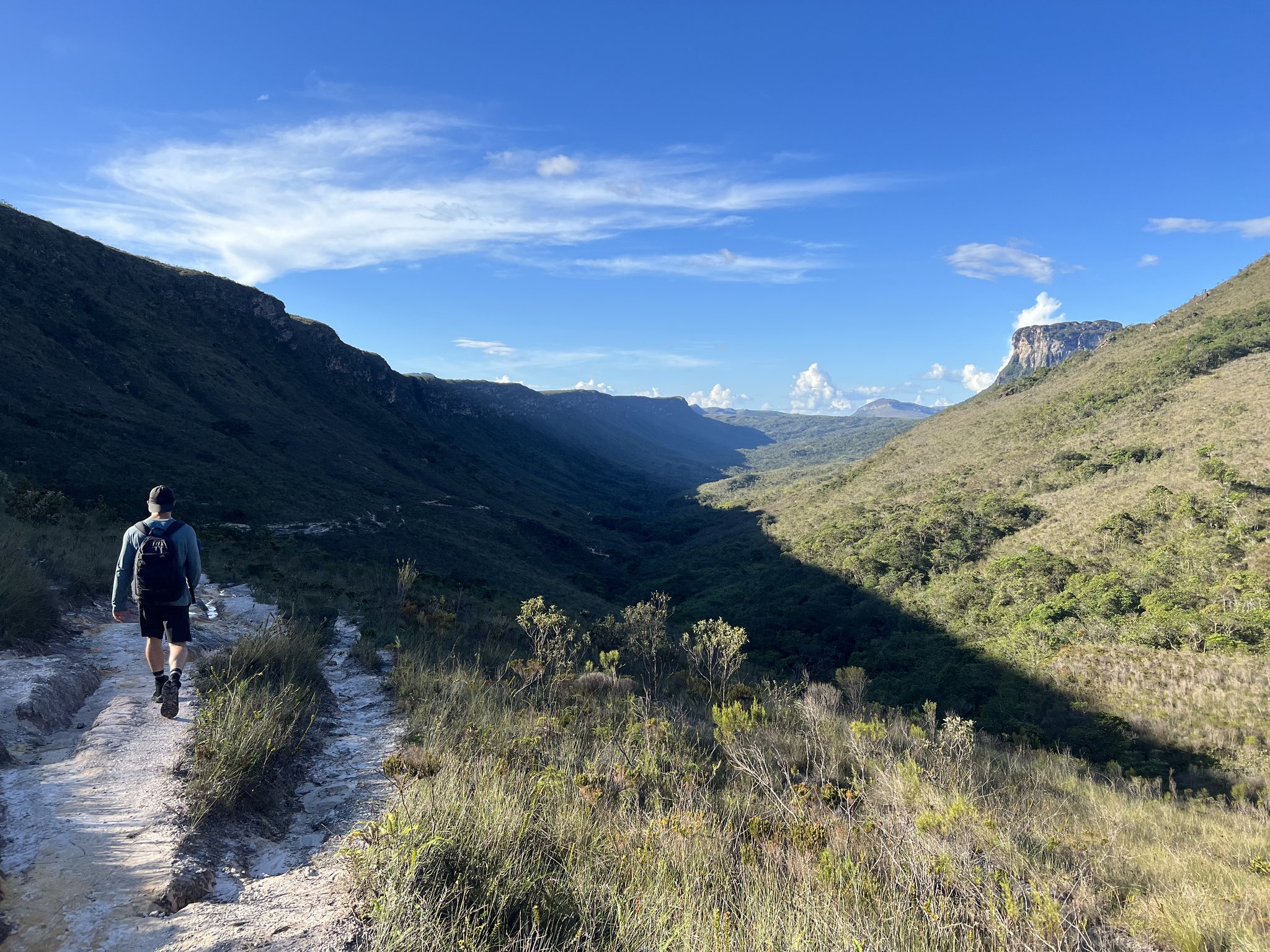 Ridge underfoot, an entire valley unfolding ahead. A solo hiker with a backpack walks a rocky ridge trail on a Chapada Diamantina trek, with a vast green valley and sandstone escarpments stretching into the distance under a bright blue sky