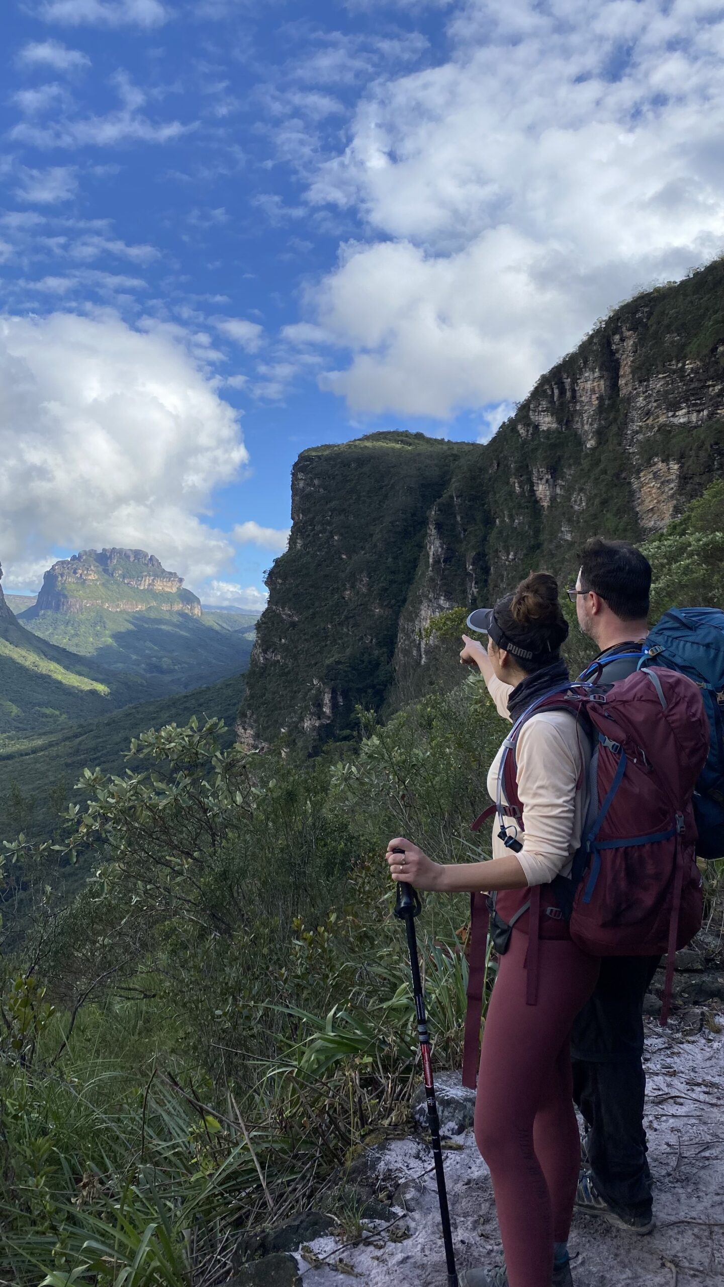 Two sets of eyes, one enormous valley — the plateau views never get old. Photo: Lucas Neves Two hikers with backpacks pause on a rocky ledge during a Chapada Diamantina trek, pointing toward a vast valley framed by towering sandstone mesas under a blue sky with scattered clouds