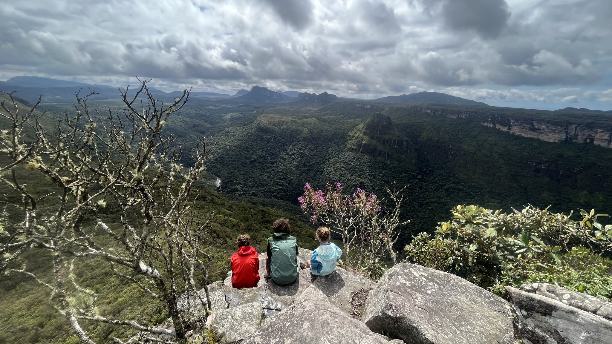 Three hikers sitting on a rocky ledge during a Chapada Diamantina trek, gazing over a vast green canyon with table-top mountains and dramatic clouds on the horizon