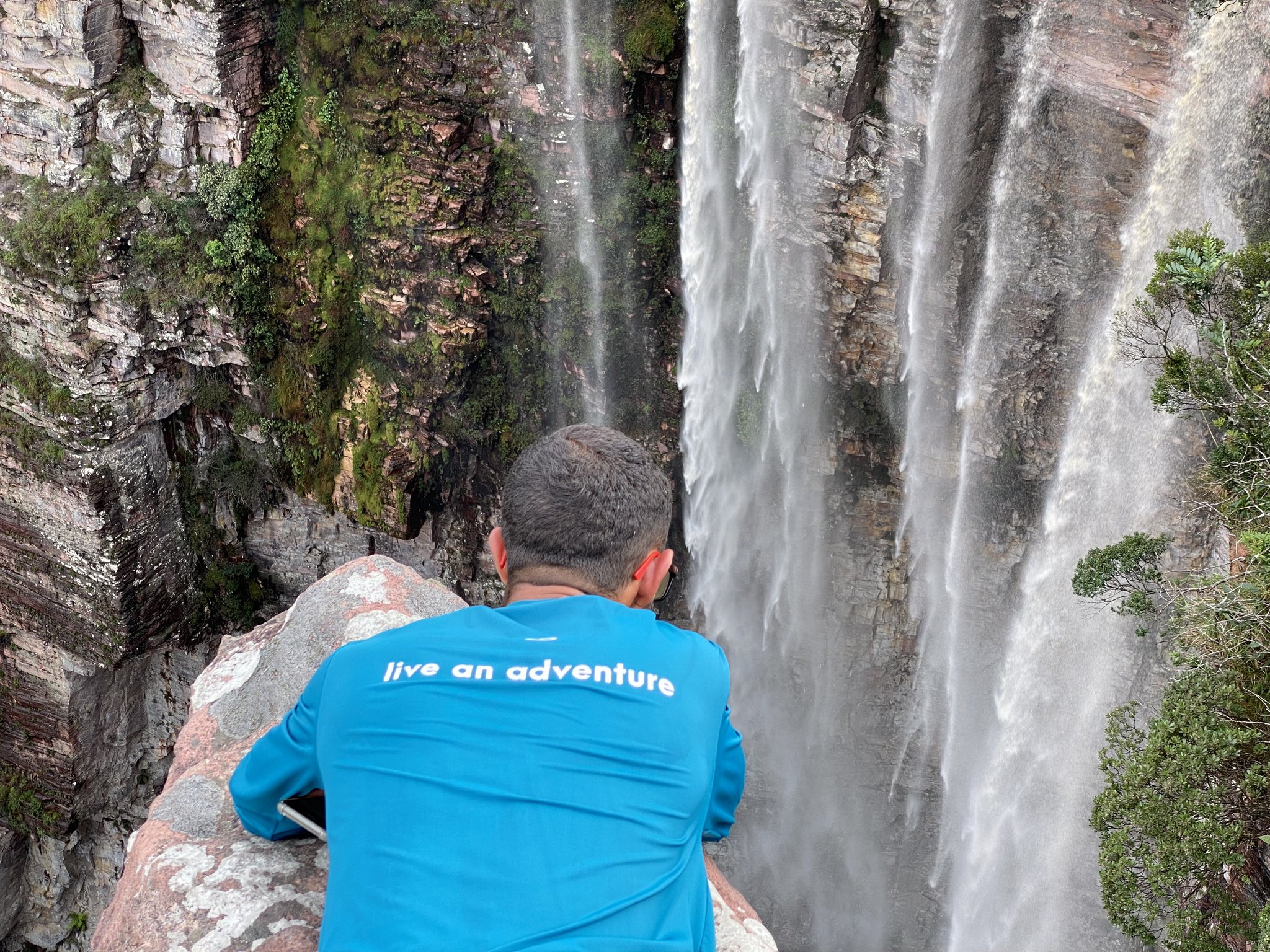 A trekker in a blue 'live an adventure' shirt leans over a rock to peer down a sheer canyon wall as a powerful waterfall cascades beside them on a Chapada Diamantina trek