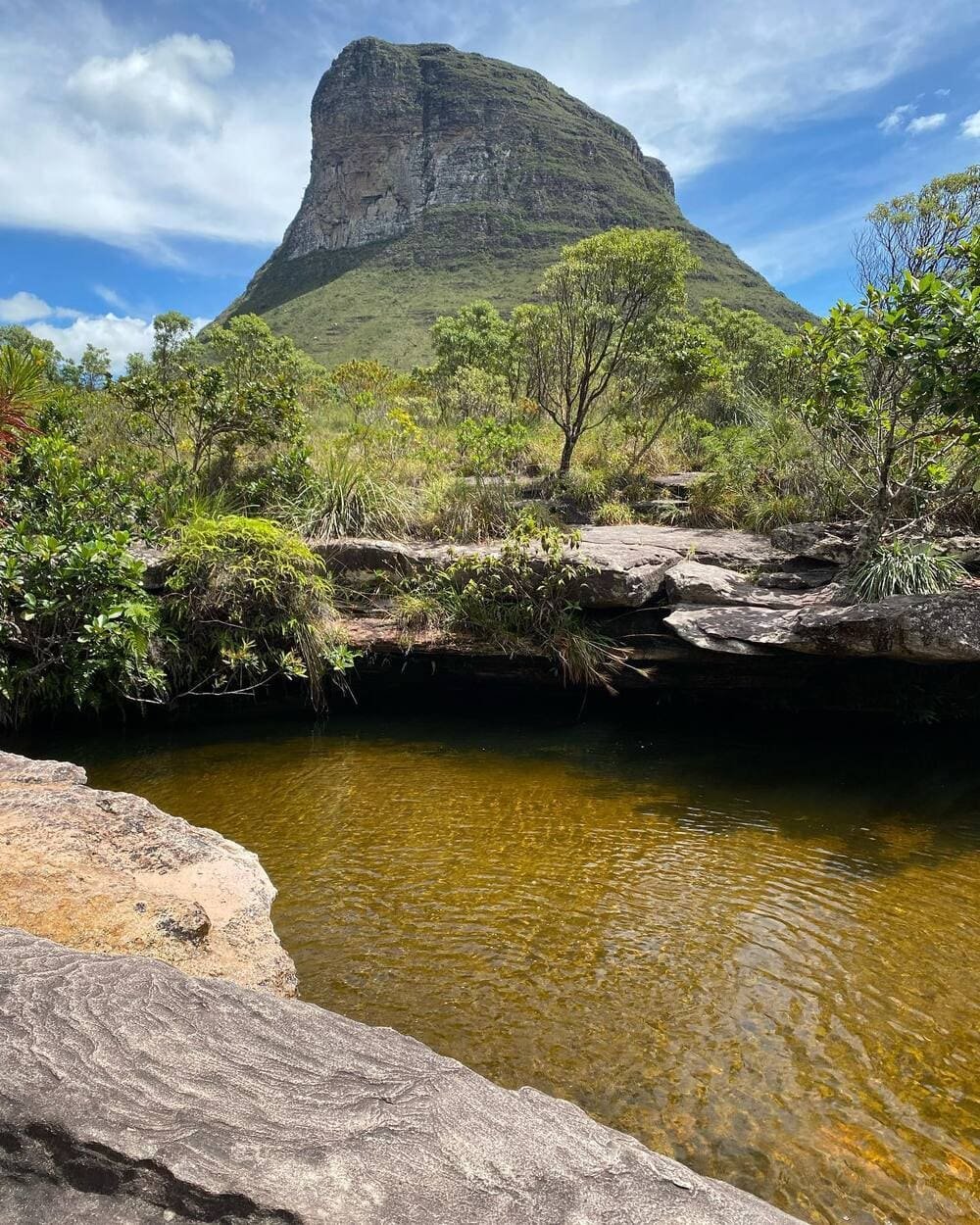 A tannin-stained natural pool reflects amber light in front of a dramatic flat-topped rocky peak rising above cerrado shrubland on a Chapada Diamantina trip