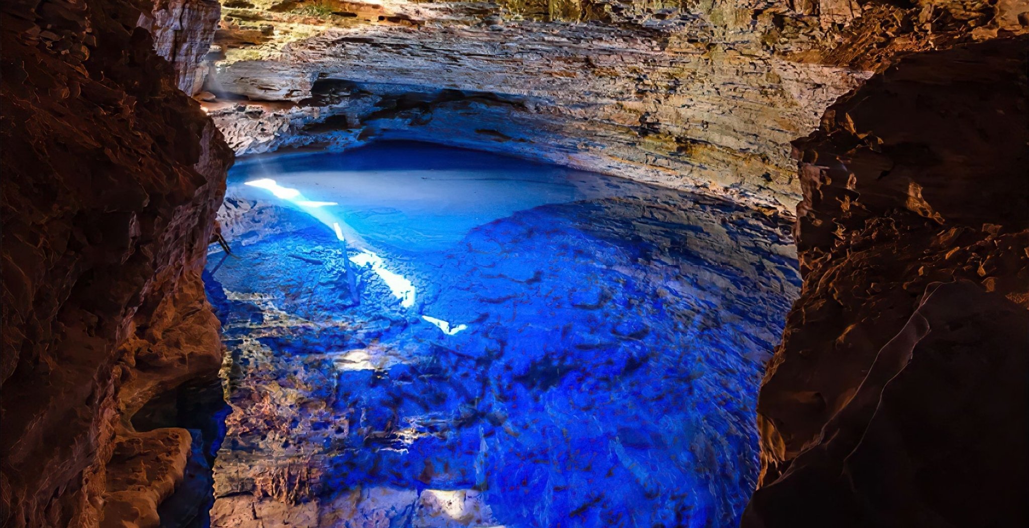 Electric-blue light floods a subterranean pool inside a sandstone cave on a Chapada Diamantina trip, with sunbeams piercing the cavern opening above