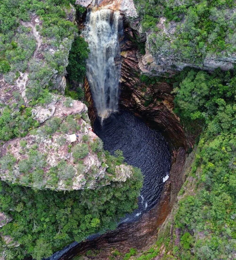 Aerial view of a tall waterfall plunging into a dark circular plunge pool inside a deep sandstone canyon on a Chapada Diamantina trip, surrounded by dense green cerrado vegetation