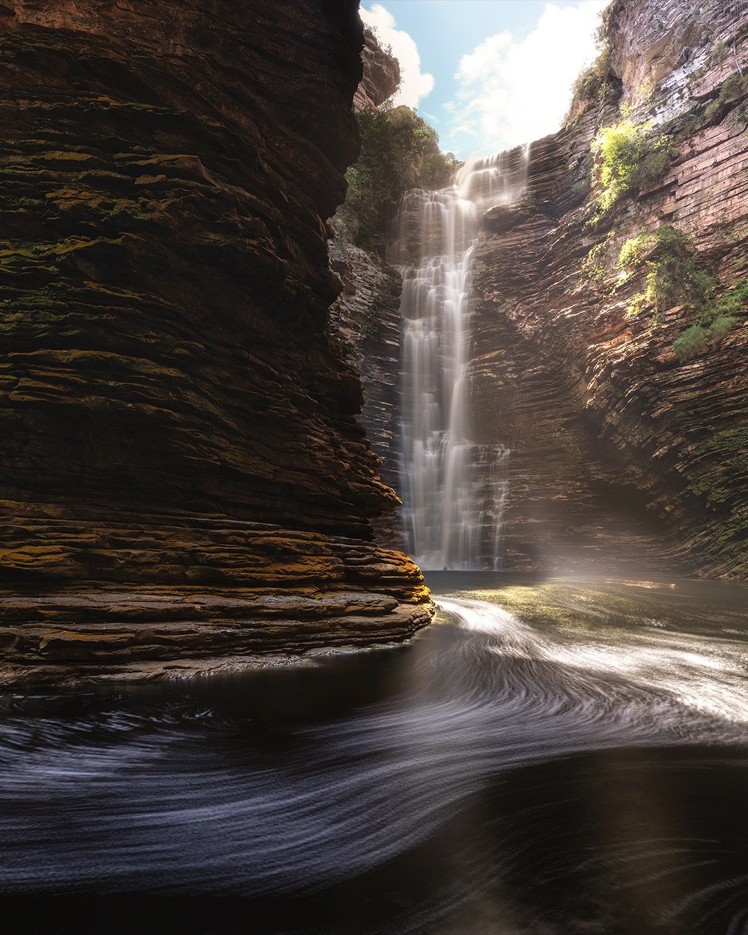 A tall waterfall cascades down layered sandstone canyon walls into a dark swirling pool on a Chapada Diamantina trip, with sunlight breaking through the narrow gorge opening above