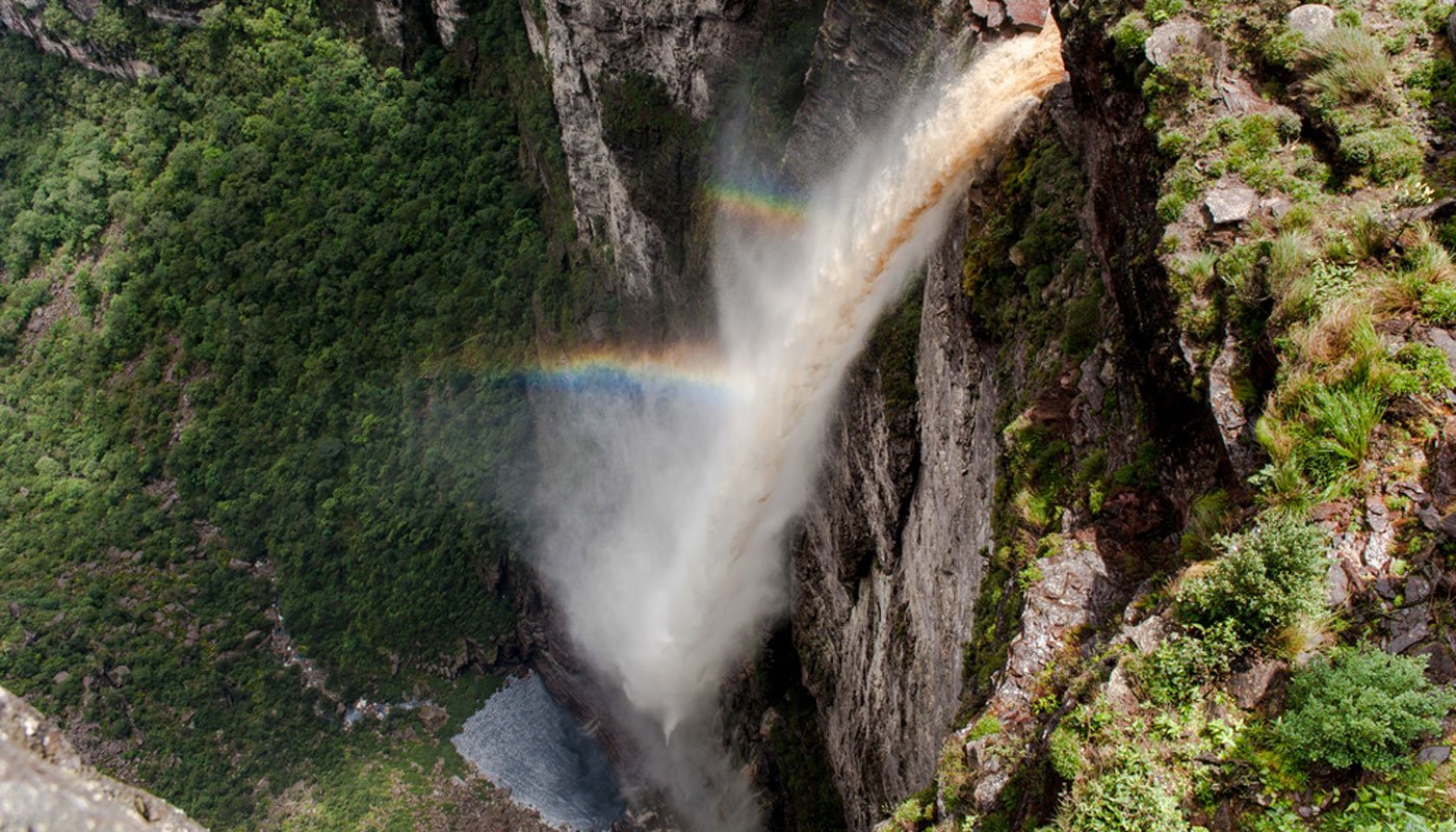 Aerial view looking straight down at a massive waterfall on a Chapada Diamantina trip, its amber-tinted torrent plunging hundreds of metres into a misty gorge with a vivid double rainbow arching through the spray and lush green cliffs on either side