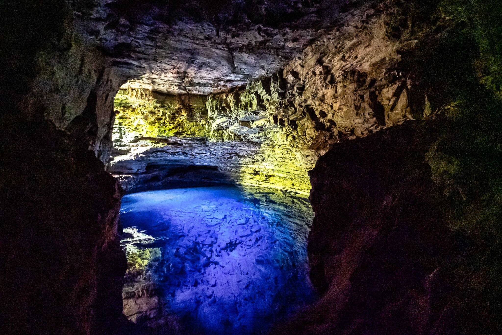 Colourful artificial lights illuminate the rocky interior of a limestone cave on a Chapada Diamantina trip, casting vivid blue and yellow hues across the subterranean pool and ancient stone walls