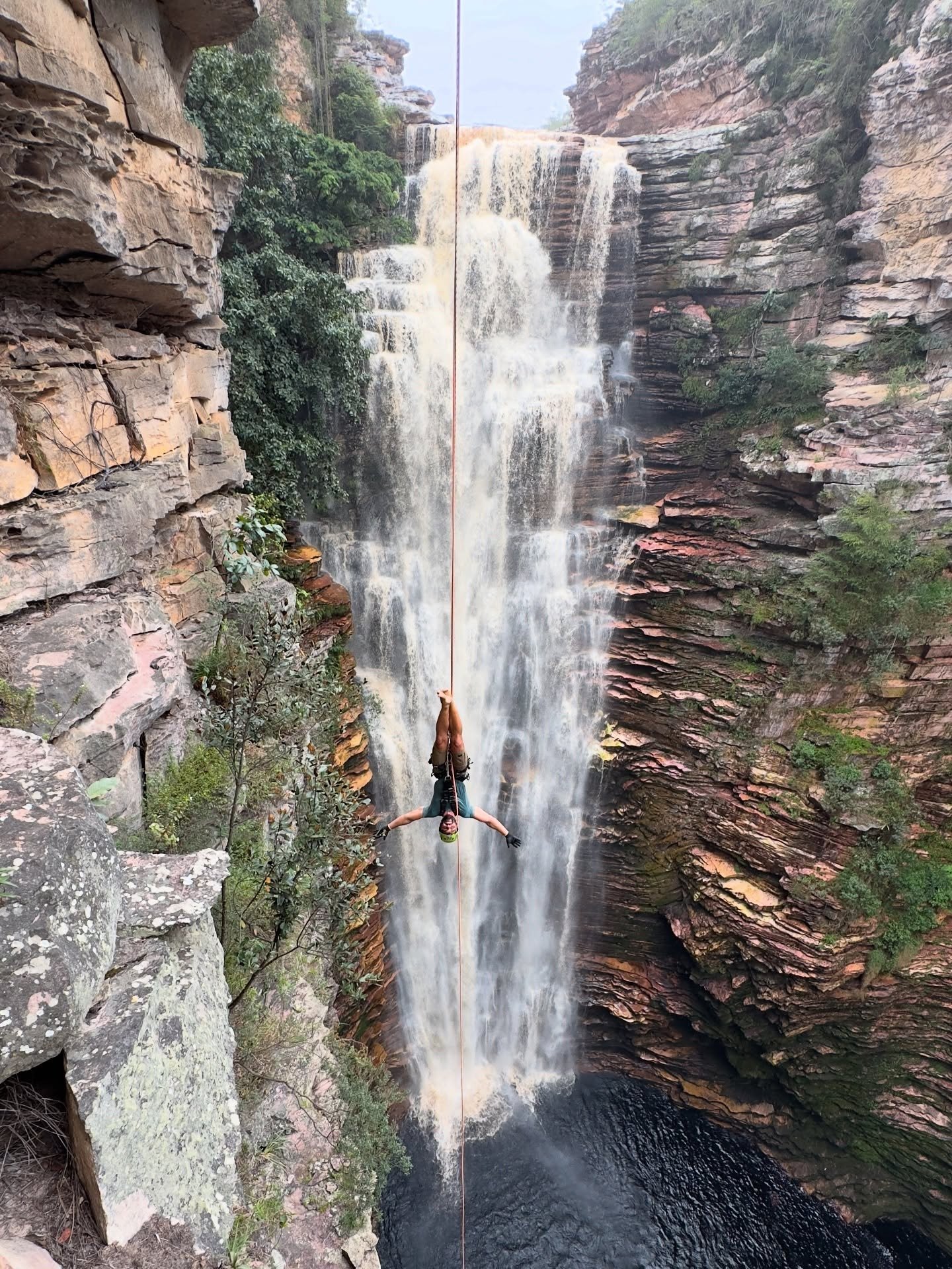 A person rappelling upside-down on a rope in front of a tall multi-tiered waterfall cascading through a deep sandstone canyon on a Chapada Diamantina trip