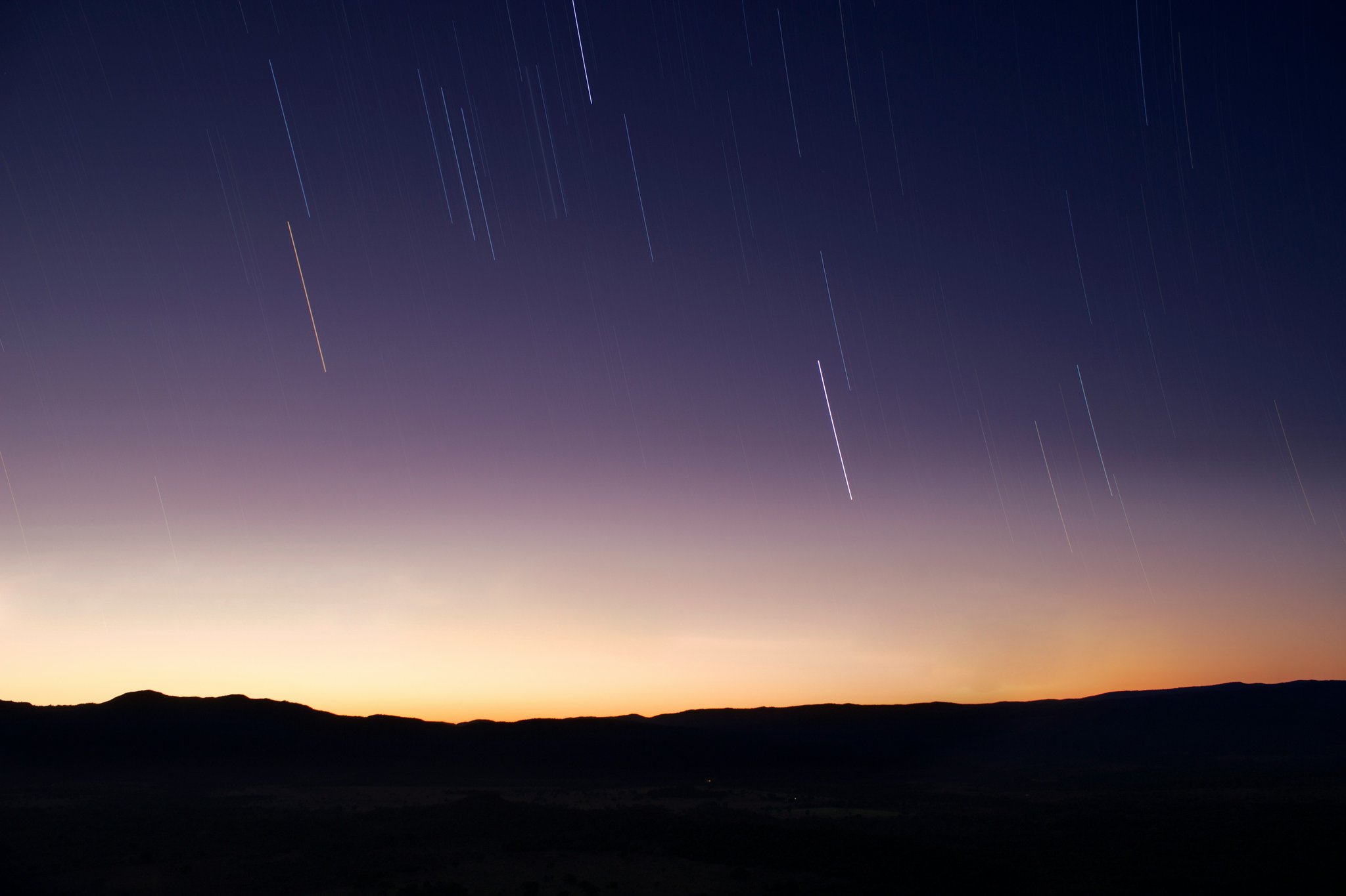 Long-exposure star trails streak across a deep purple twilight sky above the silhouetted highlands of Chapada dos Veadeiros, with a warm amber glow along the horizon