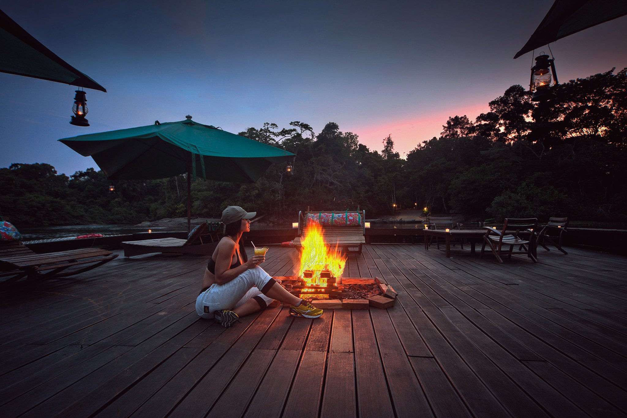 A guest sits beside an open fire on the wooden deck at Cristalino Jungle Lodge, holding a cocktail as a pink-and-blue twilight sky glows above the rainforest canopy, with hanging lanterns lighting the scene