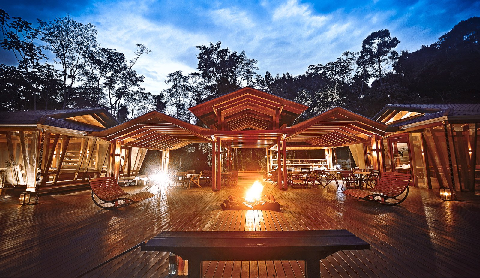 The open-air timber deck at Cristalino Jungle Lodge glows at dusk, with a crackling bonfire at its centre, rocking chairs on either side, a bar visible in the background, and tall rainforest trees silhouetted against a deep blue twilight sky