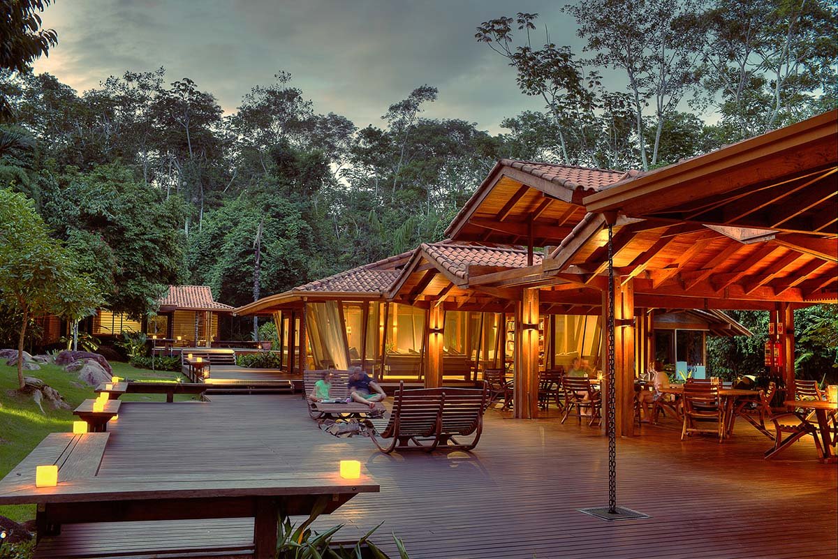 Cristalino Jungle Lodge's timber open-air deck glowing with candles and warm lamp light at dusk, with guests relaxing on wooden rocking chairs and dense rainforest rising in the background