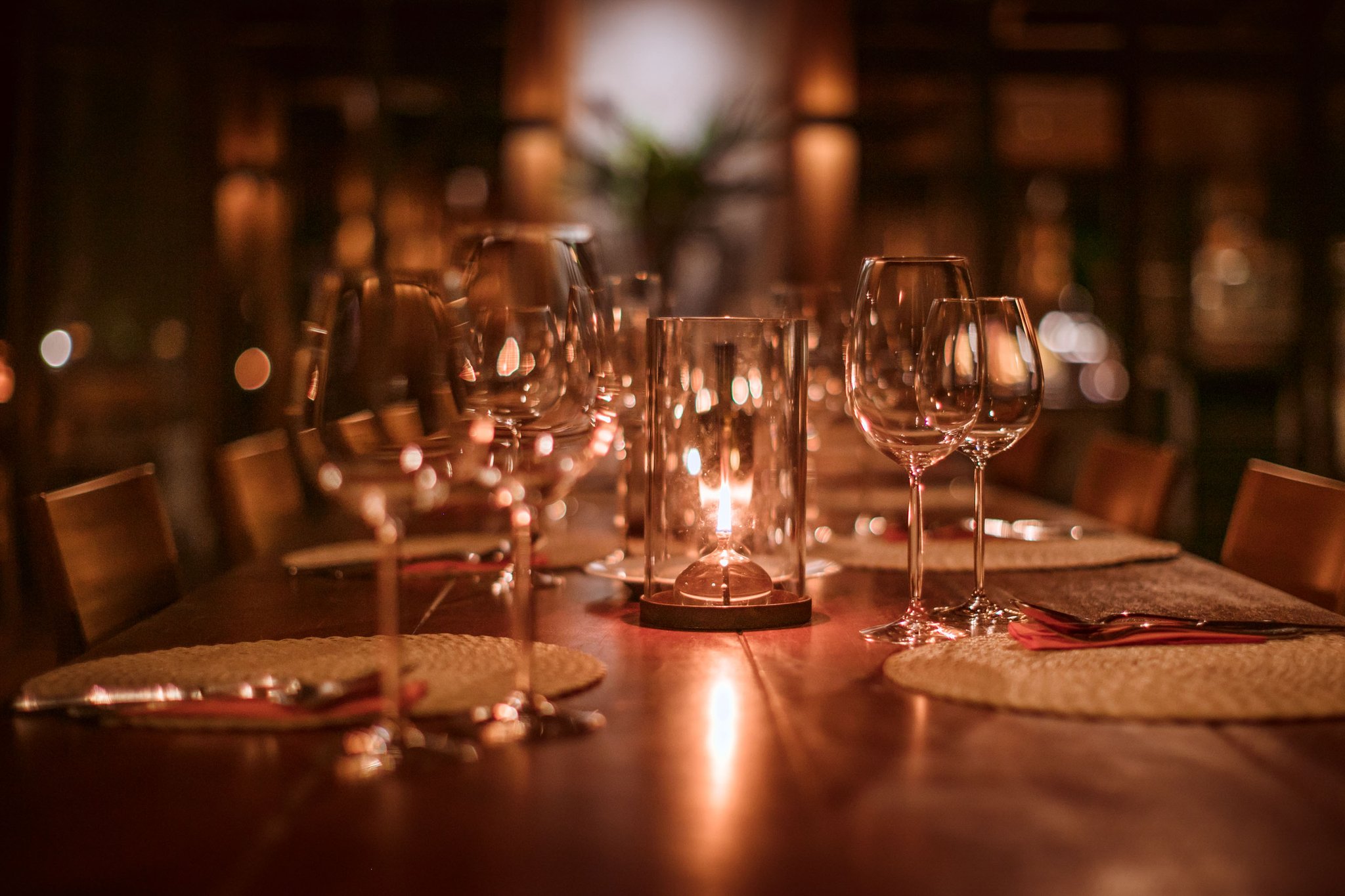 A long polished wooden dining table set for guests at Cristalino Jungle Lodge, with crystal wine glasses, woven placemats, red napkins, and a glowing glass candle lantern casting warm amber light across the room