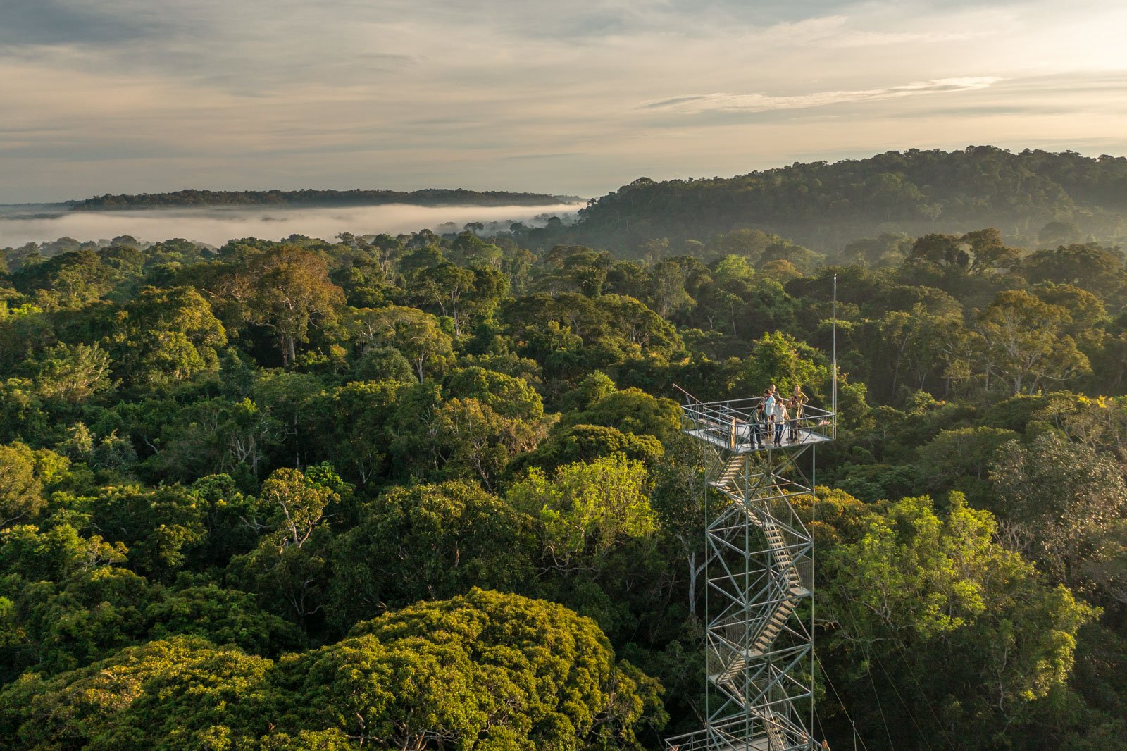 Visitors stand on a tall metal observation tower rising above the Amazon rainforest canopy at Cristalino Jungle Lodge, with low morning mist drifting over the treetops and forested hills in the background