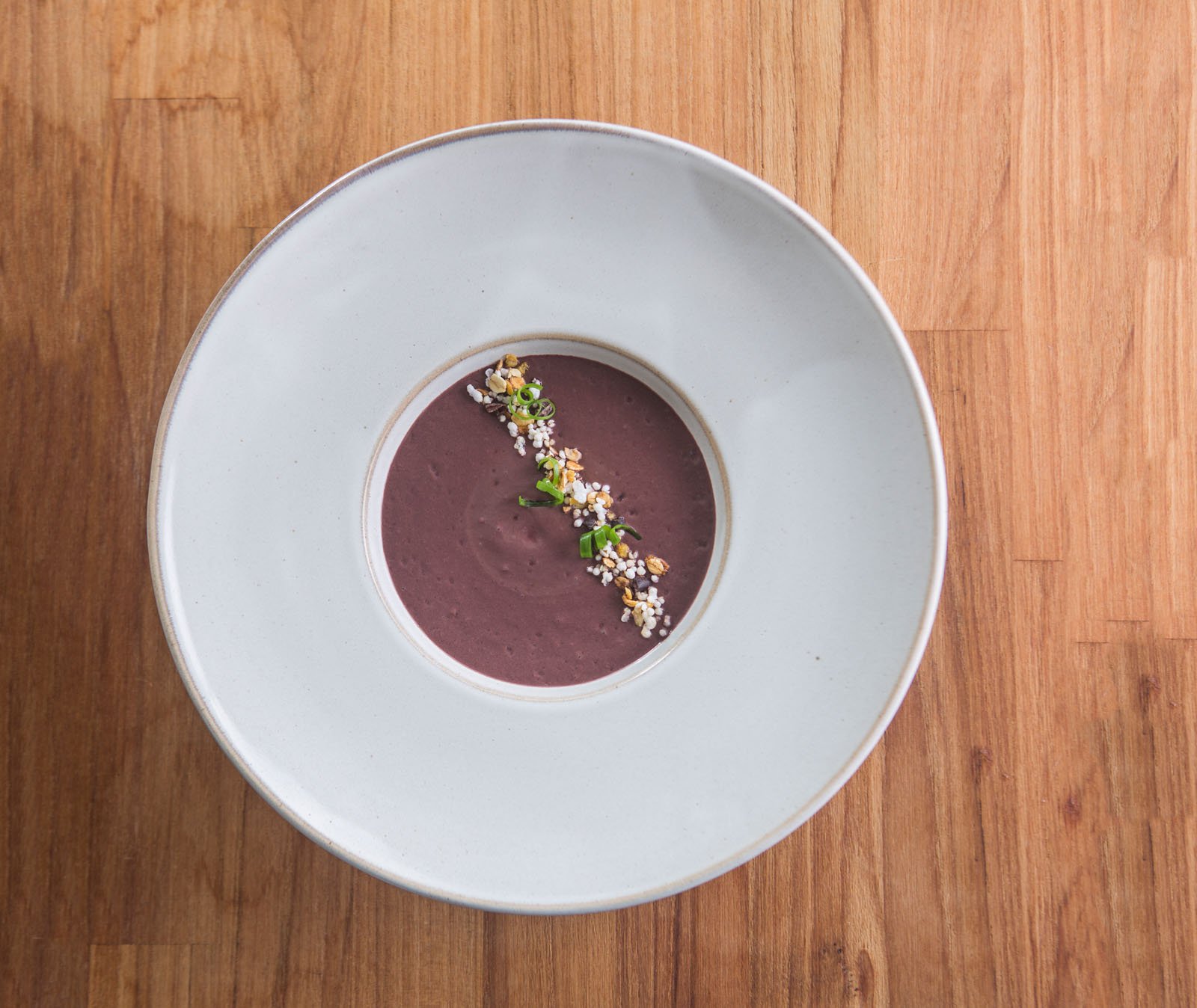 A smooth deep-purple cream soup served in a wide white ceramic bowl at Cristalino Jungle Lodge, garnished with a diagonal line of toasted grains, puffed seeds, and sliced spring onion