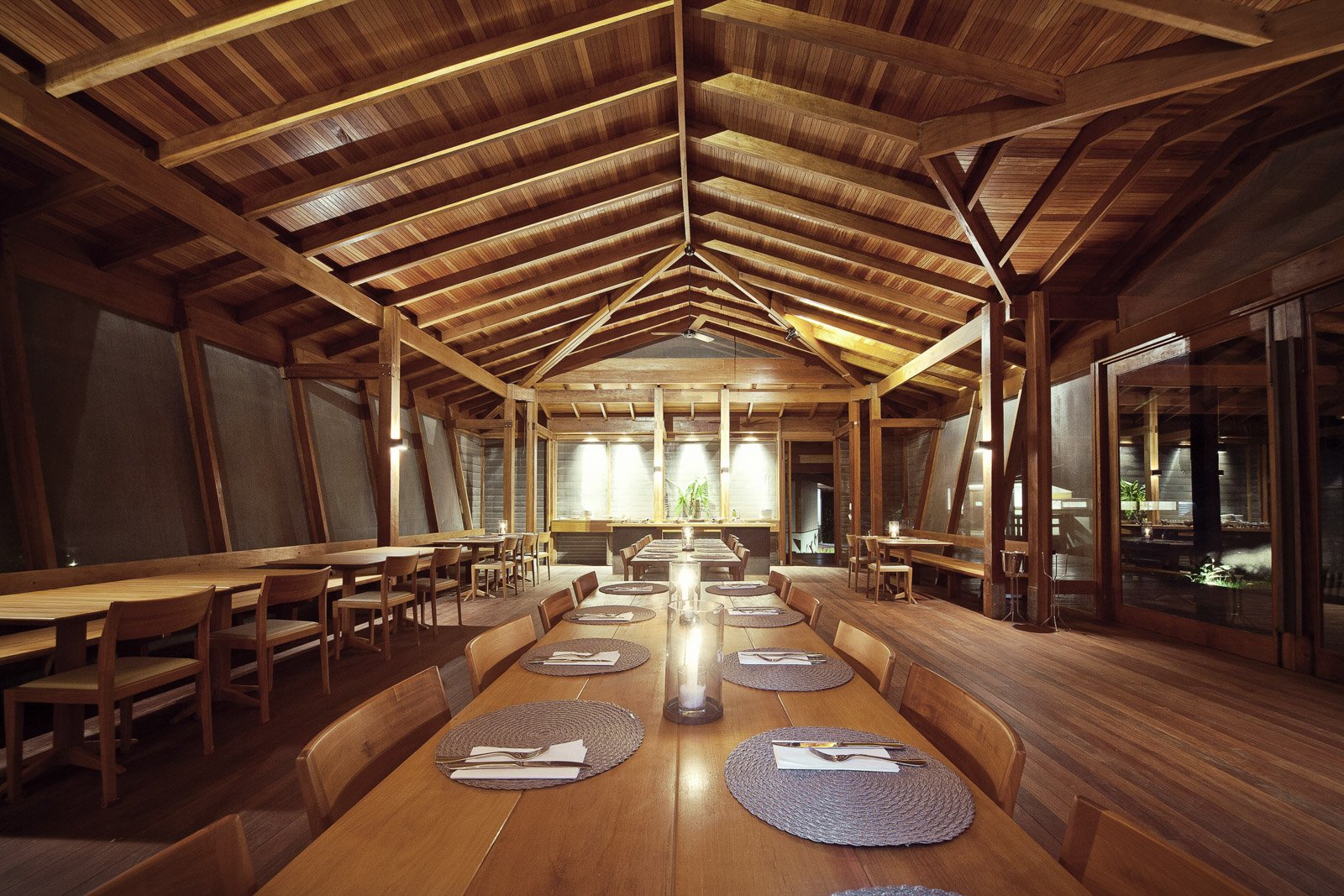 The Cristalino Jungle Lodge restaurant dining room with a soaring vaulted timber ceiling, long communal wooden tables set with woven placemats and candles, and warm pendant lighting