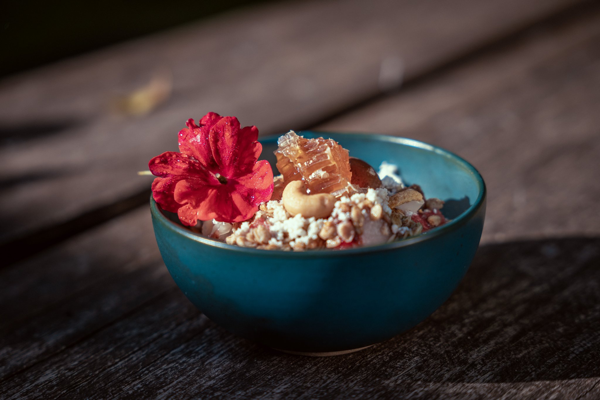 A teal ceramic bowl at Cristalino Jungle Lodge filled with granola, nuts, honeycomb, and fresh fruit, garnished with a vivid red edible flower, served on a weathered wooden surface