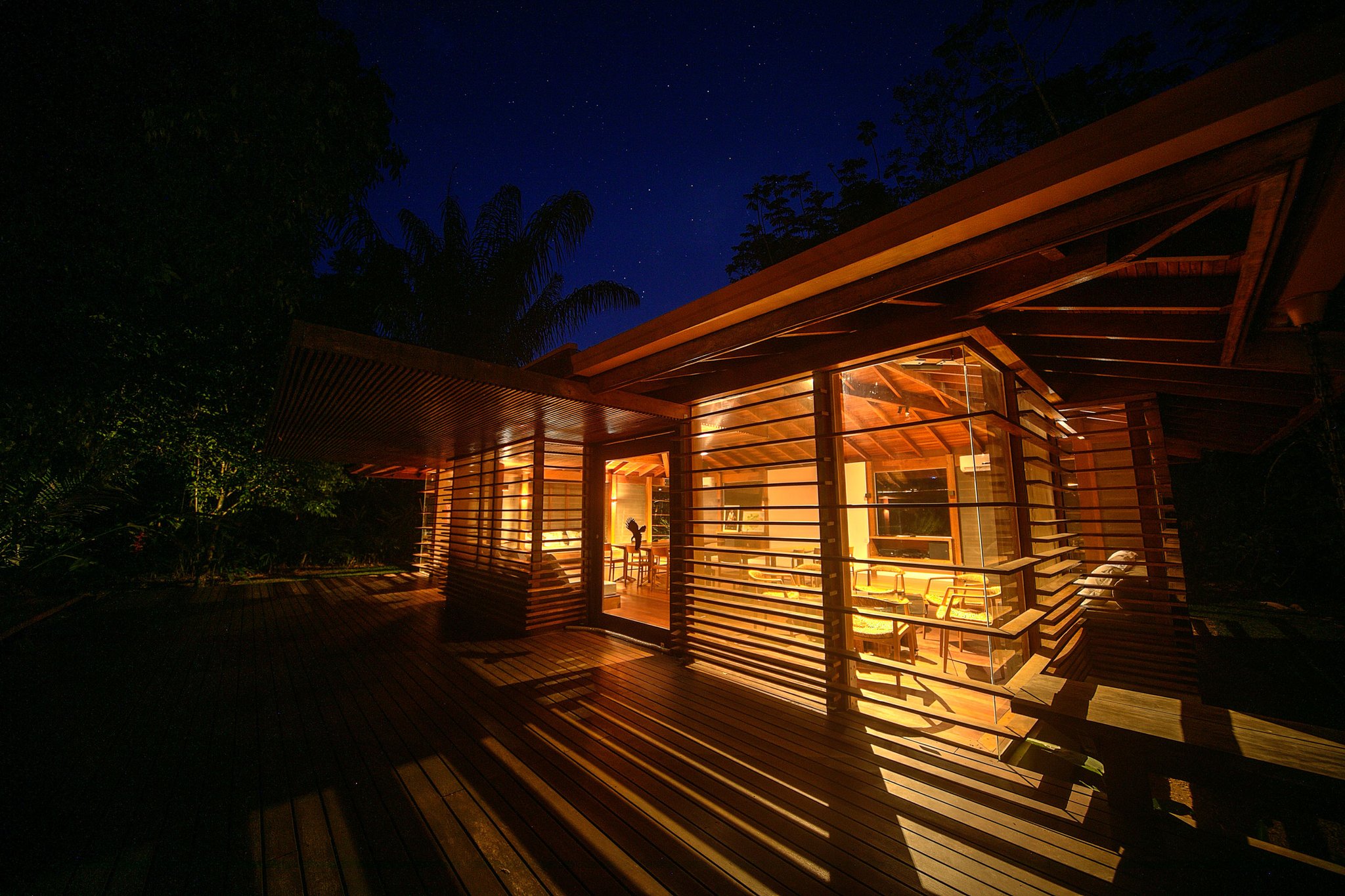 Cristalino Jungle Lodge glowing from within at night, its timber-slatted walls and wide wooden deck illuminated by warm amber light beneath a star-filled sky