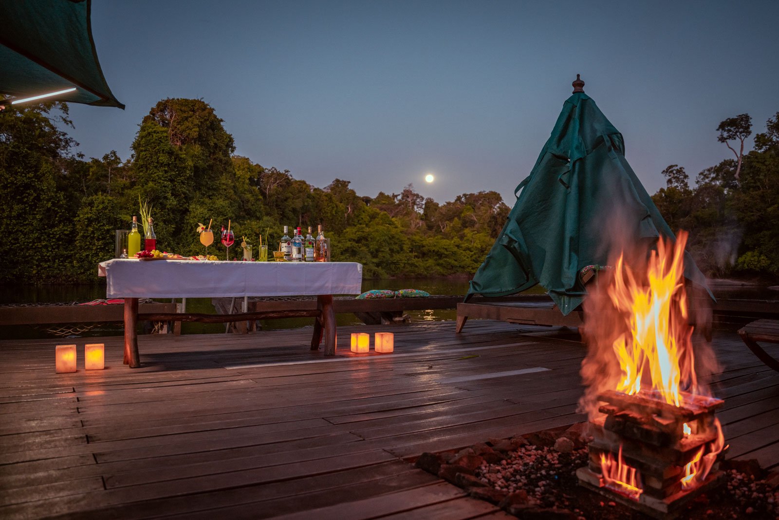 An open timber deck at Cristalino Jungle Lodge at dusk, with a roaring bonfire in the foreground, paper lantern candles lining the floor, a white-clothed cocktail table set with colorful drinks and bottles, and a full moon rising above the rainforest treeline