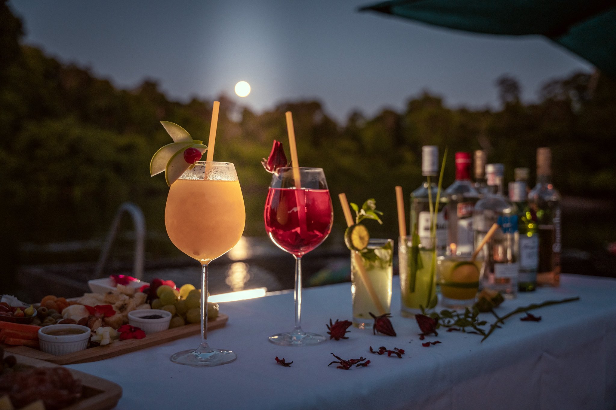 Four garnished cocktails and a charcuterie board on a white-draped table at Cristalino Jungle Lodge, with a full moon rising over the rainforest canopy in the background
