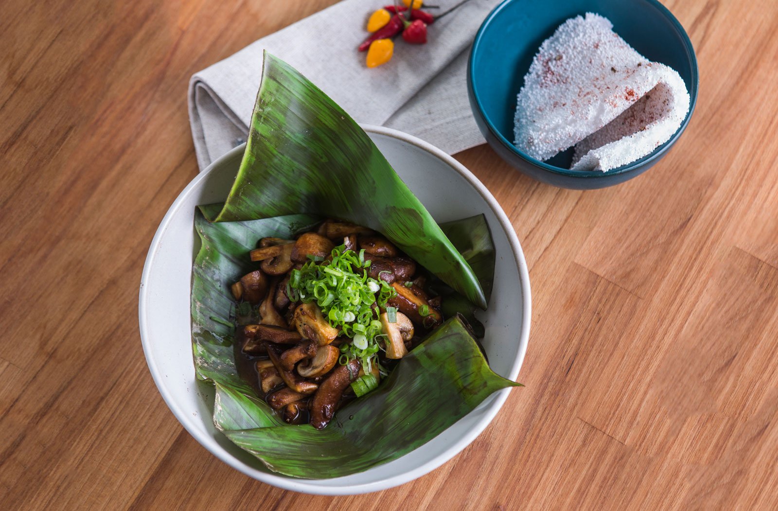Sautéed mushrooms garnished with sliced spring onions, served in a banana leaf inside a white ceramic bowl at Cristalino Jungle Lodge, with a side of cassava flatbread dusted in red spice and small chilli peppers on a linen napkin