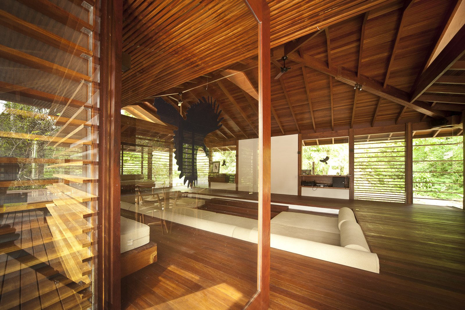Open-air common lounge at Cristalino Jungle Lodge featuring warm hardwood floors, a vaulted timber ceiling, white day-bed sofas, and louvred walls opening onto lush green rainforest
