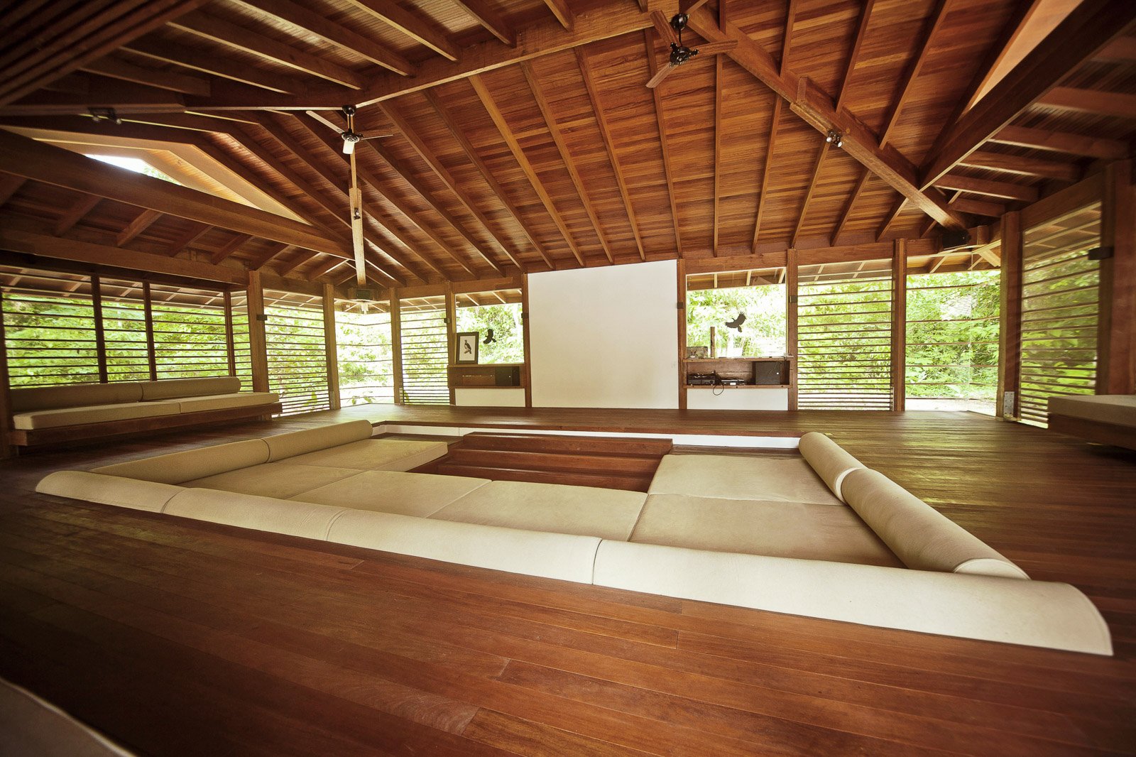 The open-air lounge at Cristalino Jungle Lodge features a sunken seating area with cream cushions, polished hardwood floors, a large projector screen, and a vaulted timber roof with ceiling fans, surrounded by lush green forest on all sides.