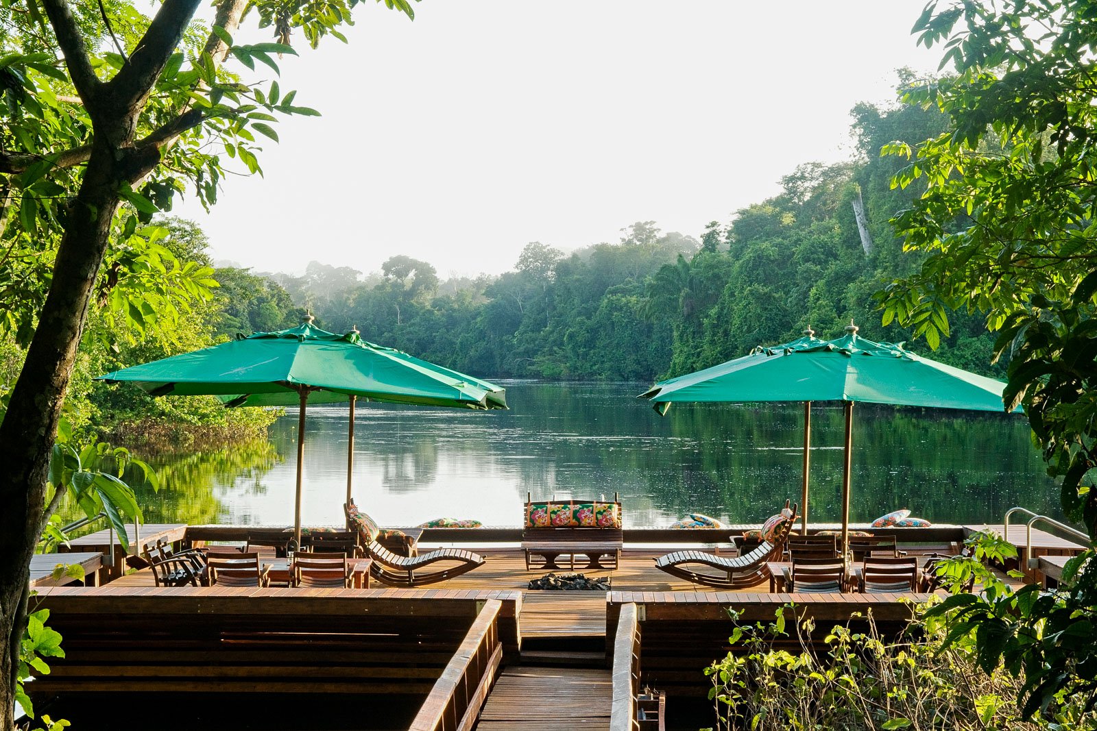 A wooden deck terrace at Cristalino Jungle Lodge with teak sun loungers and green parasols overlooking a calm jungle river flanked by dense rainforest