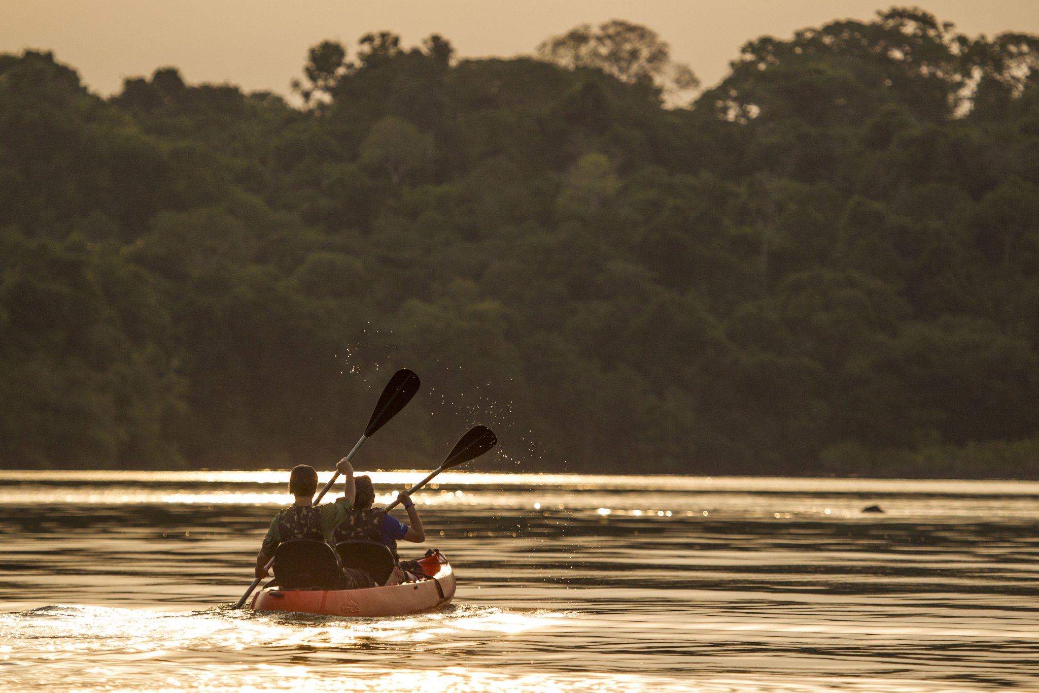 Two paddlers in a tandem kayak glide across a glassy river at golden hour, with dense rainforest canopy reflected on the water at Cristalino Jungle Lodge