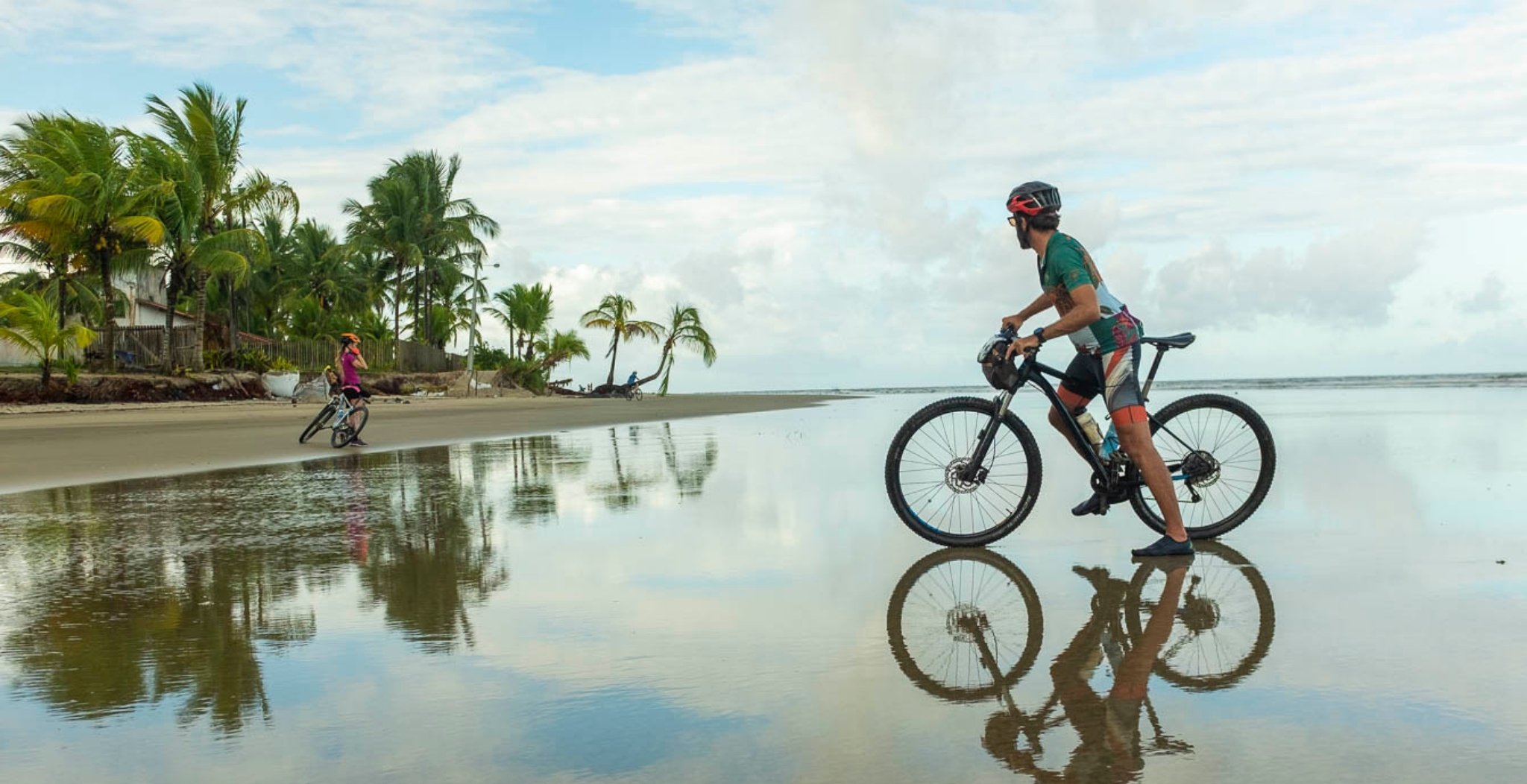 Three cyclists riding mountain bikes along a mirror-flat wet beach lined with palm trees near Itacaré during a cycling Bahia tour